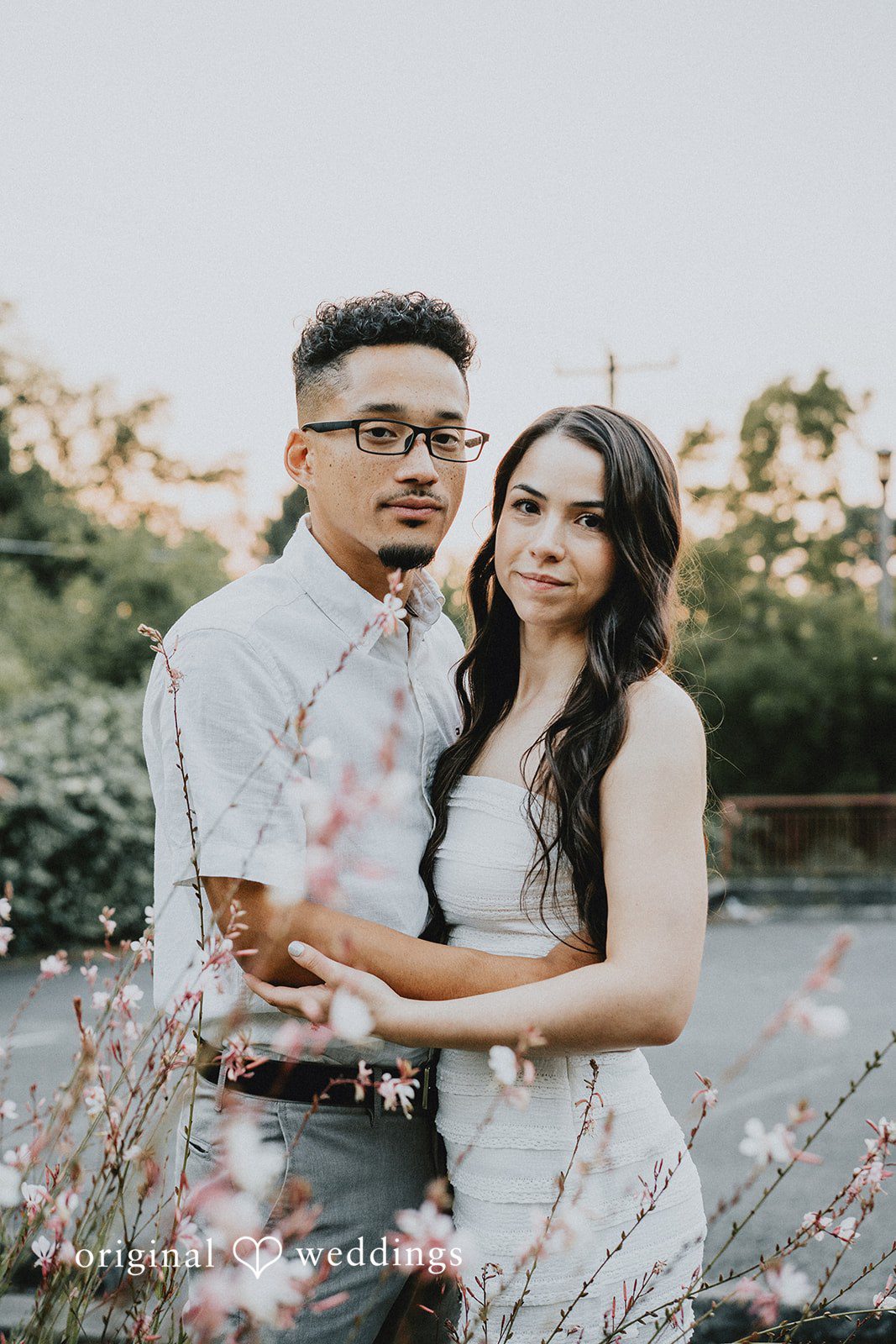 Couple standing close together and smiling against a backdrop of foliage and soft lighting.