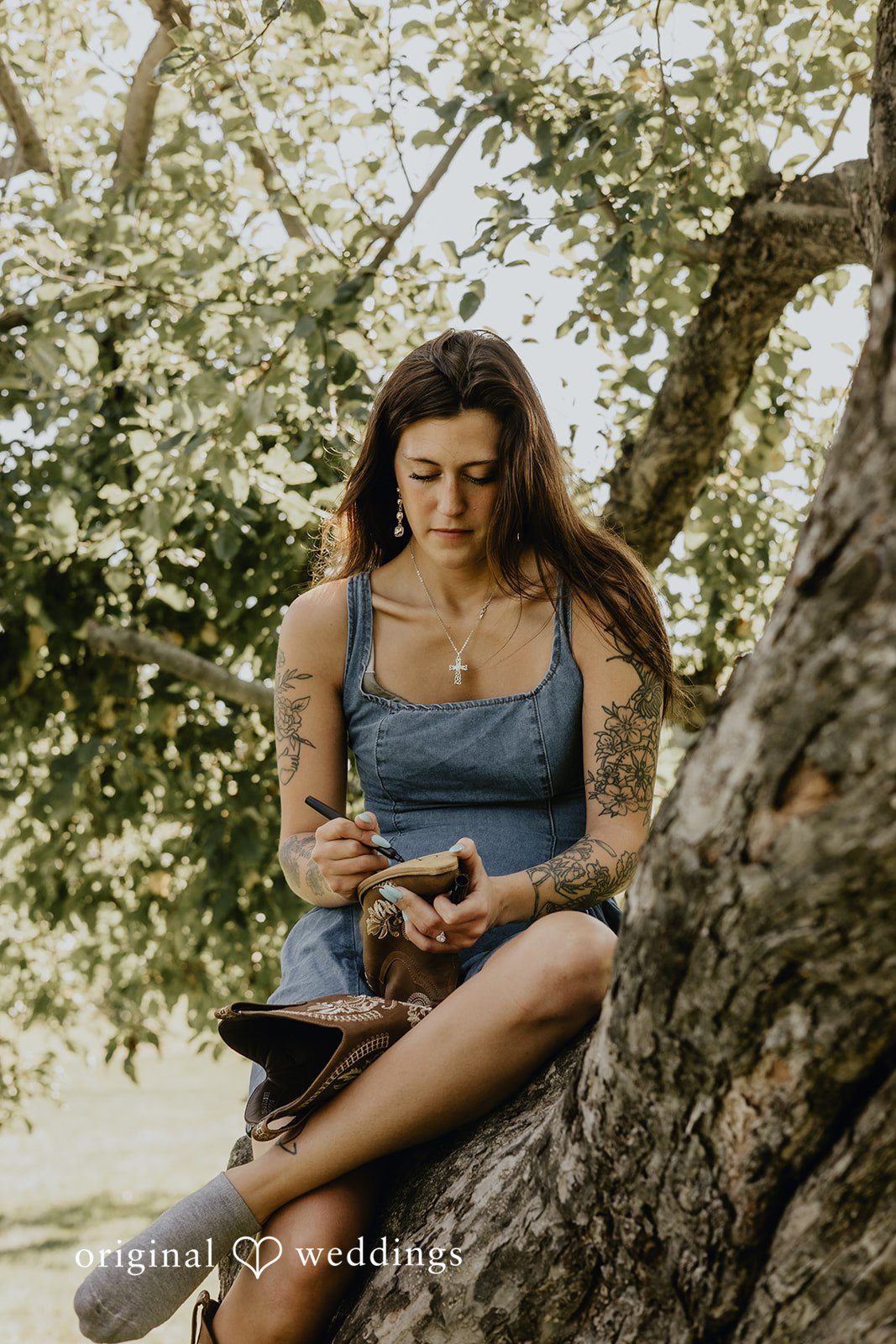 Meghan + Kal A woman with arm tattoos sits in a large tree writing on the sole of a cowboy boot.