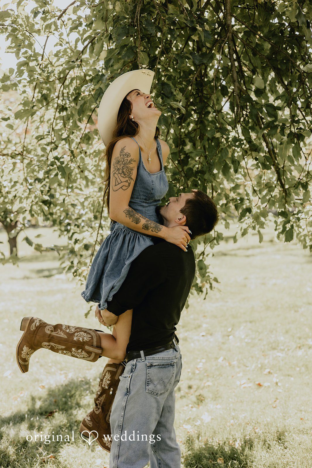 Meghan + Kal A man lifts a woman up in the air near a large tree.