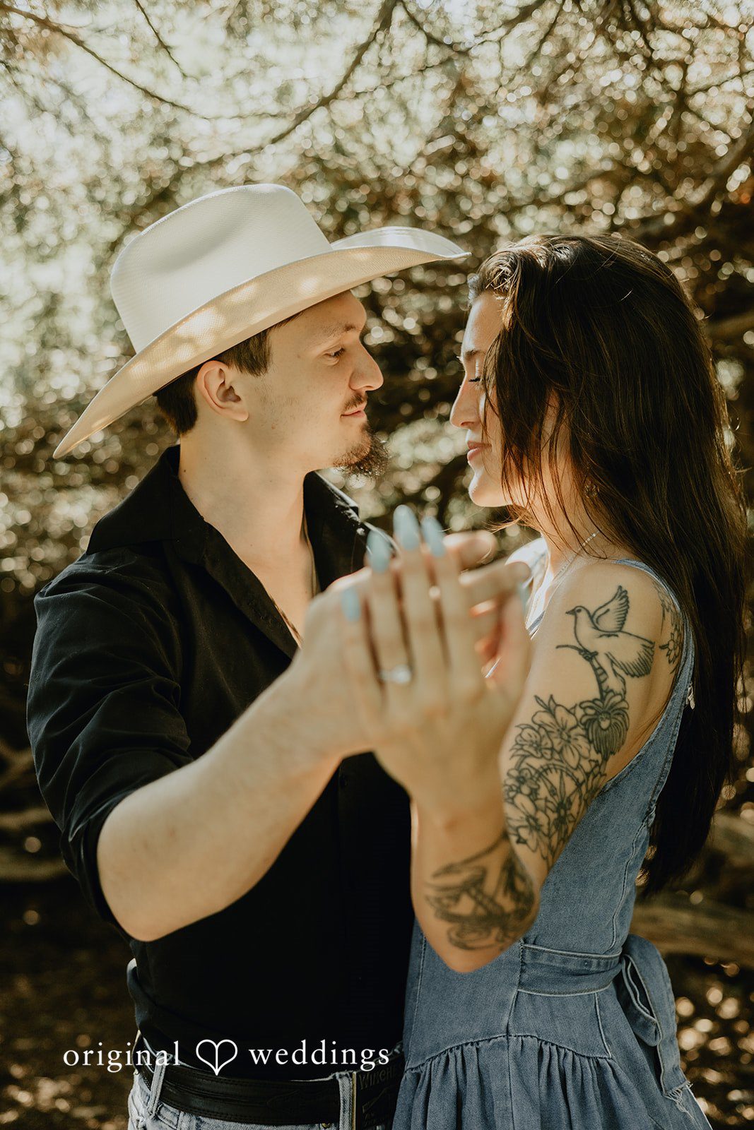 Meghan + Kal A man wearing a cowboy hat holds a woman close while facing each other.