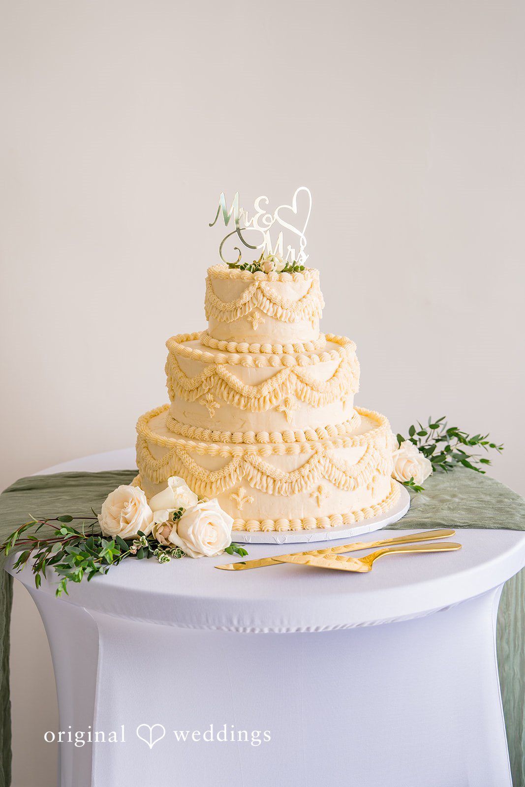 Kalina + Malakai A three-tiered white wedding cake with subtle gold leaf detailing on a table.
