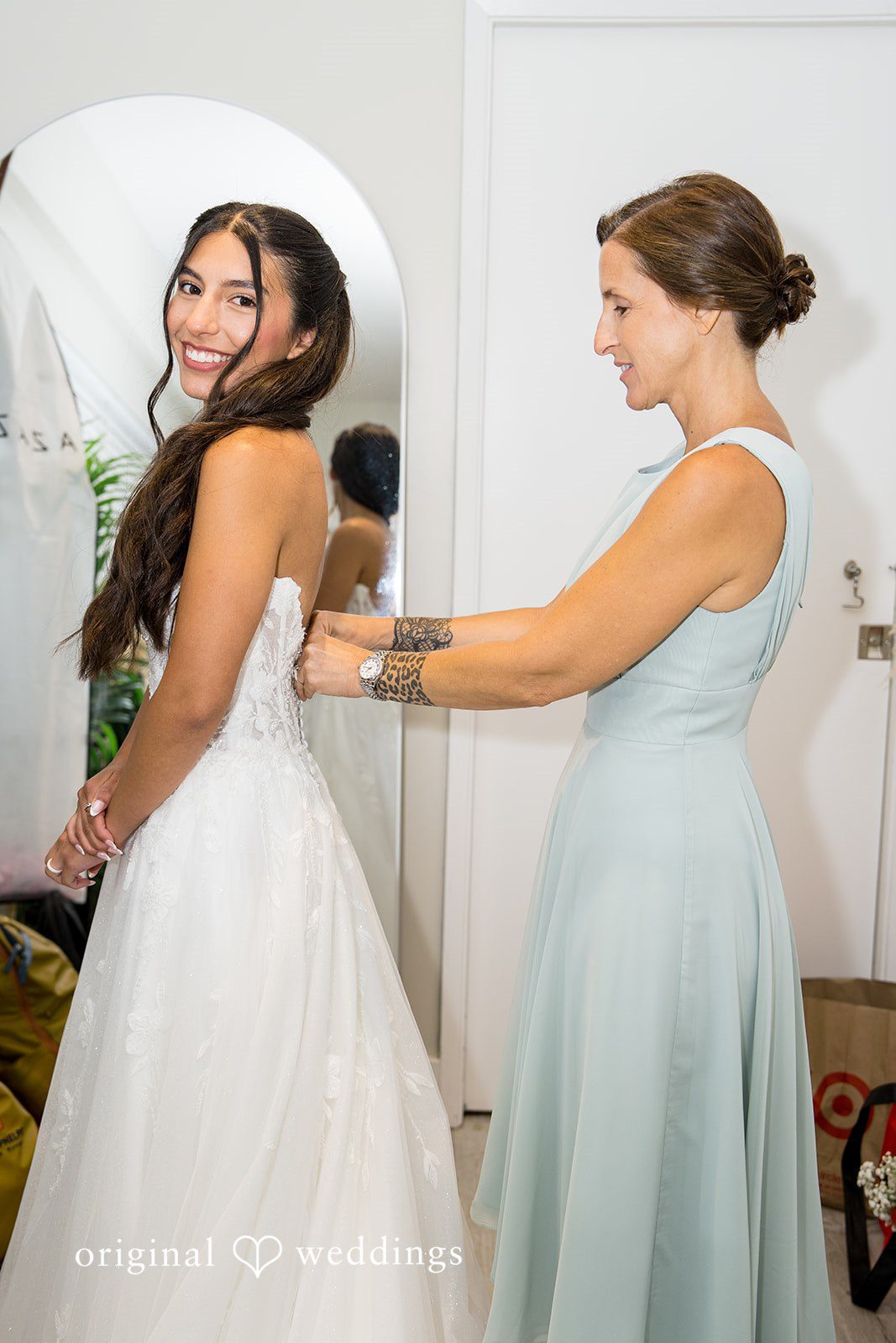 Kalina + Malakai A bridesmaid in a sage green dress helping the bride adjust her wedding gown.