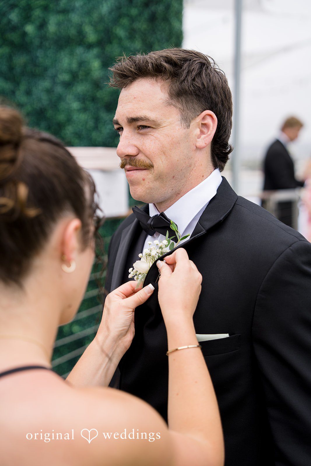 Kalina + Malakai A woman pinning a white floral boutonniere onto the lapel of a groom's black tuxedo jacket