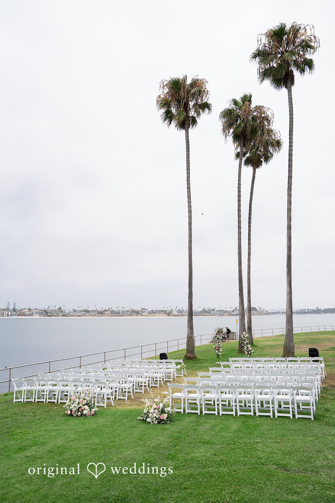 Kalina + Malakai White ceremony chairs arranged on a lawn overlooking the water with tall palm trees.