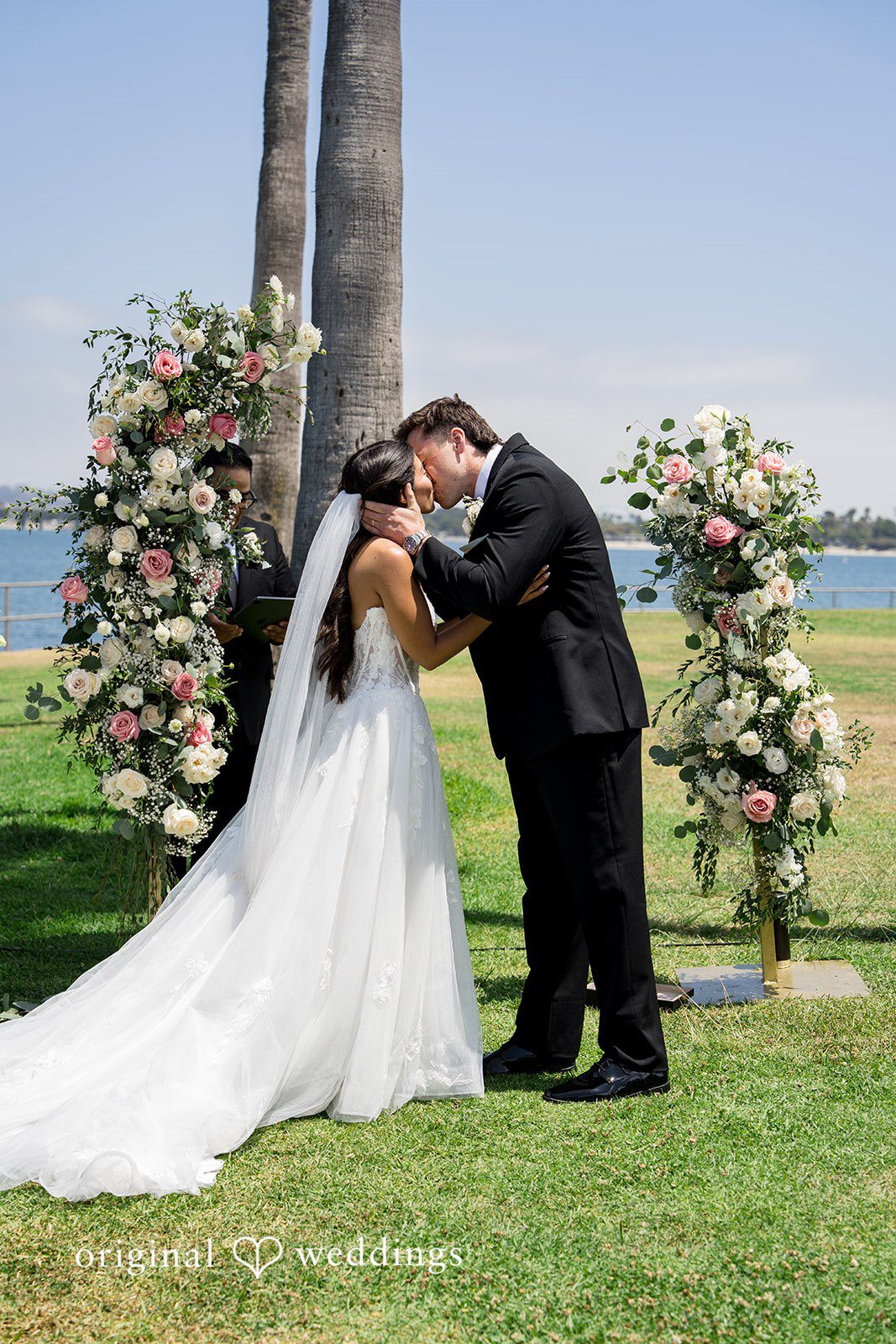 Kalina + Malakai Bride and groom kissing beneath a floral archway by the waterfront.