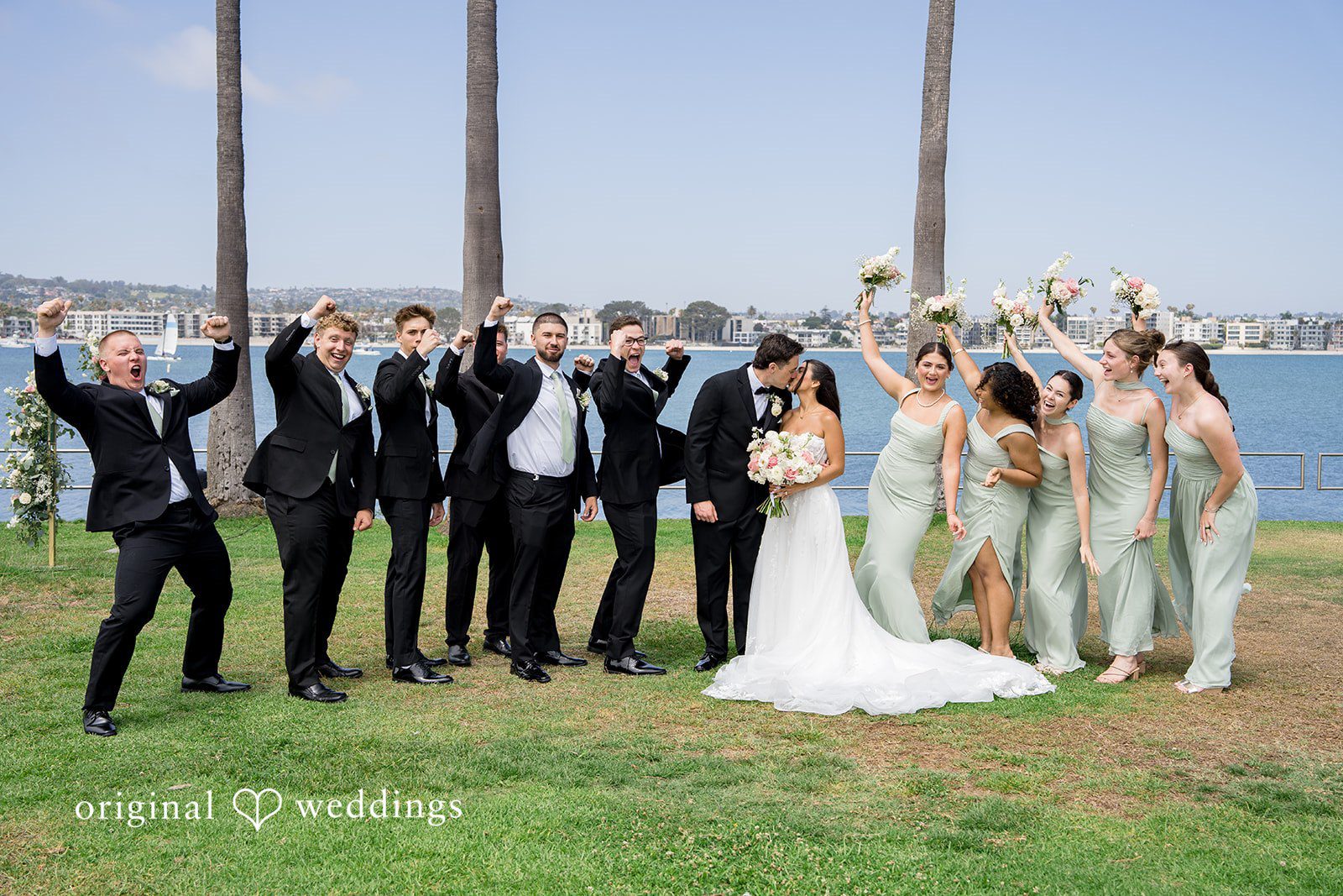 Kalina + Malakai Full wedding party cheering with raised hands on a lawn near the water.
