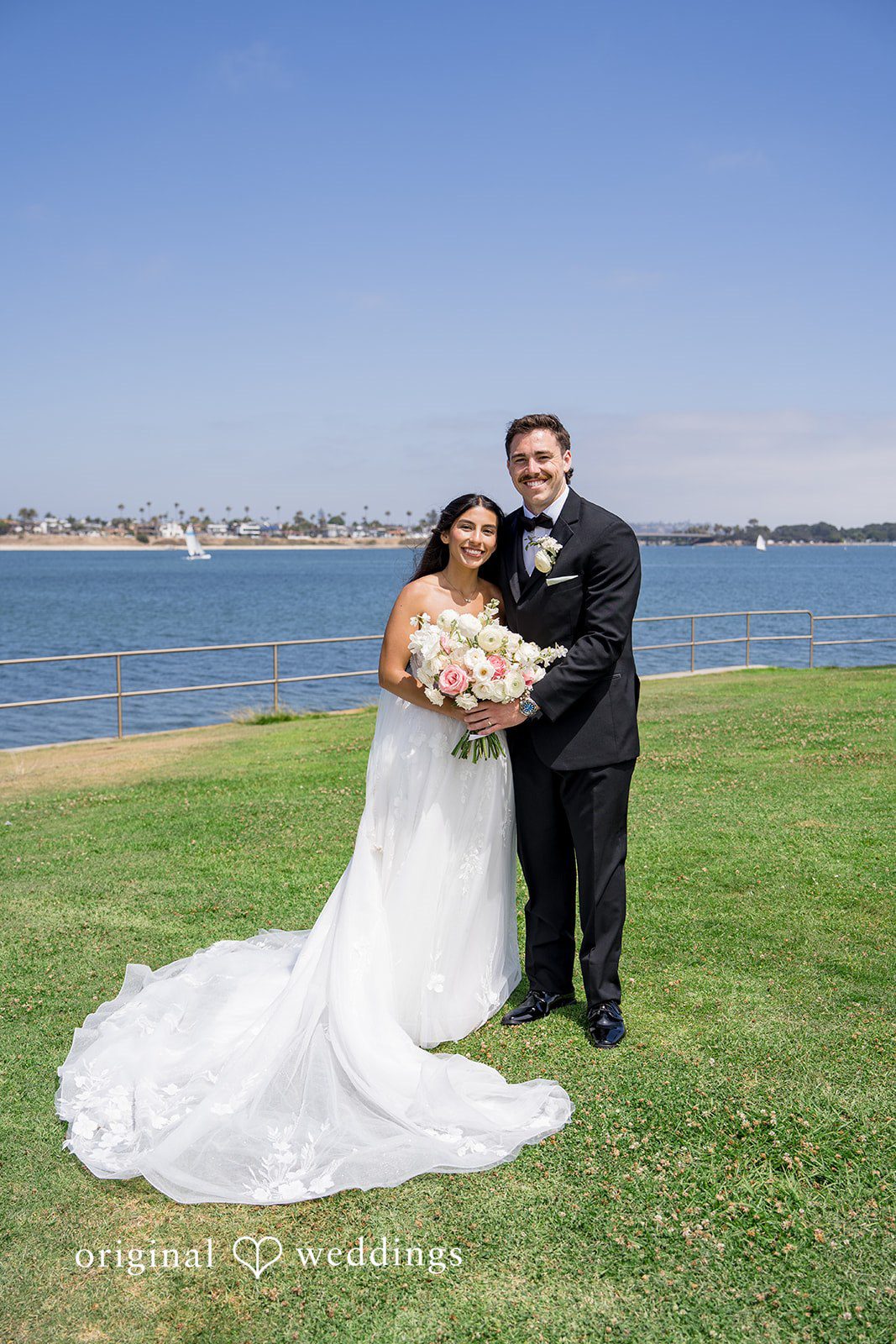 Kalina + Malakai Bride and groom embracing on a bright green lawn near the water.