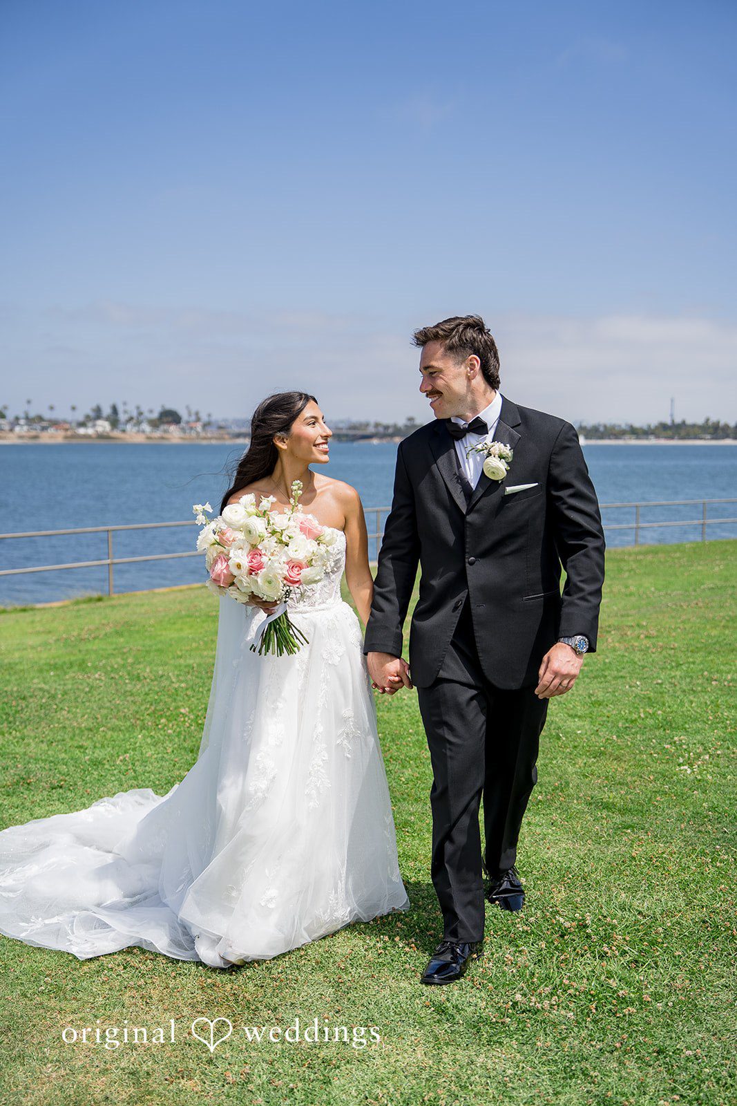 Kalina + Malakai Bride and groom walking hand-in-hand across a grassy field near a bay.