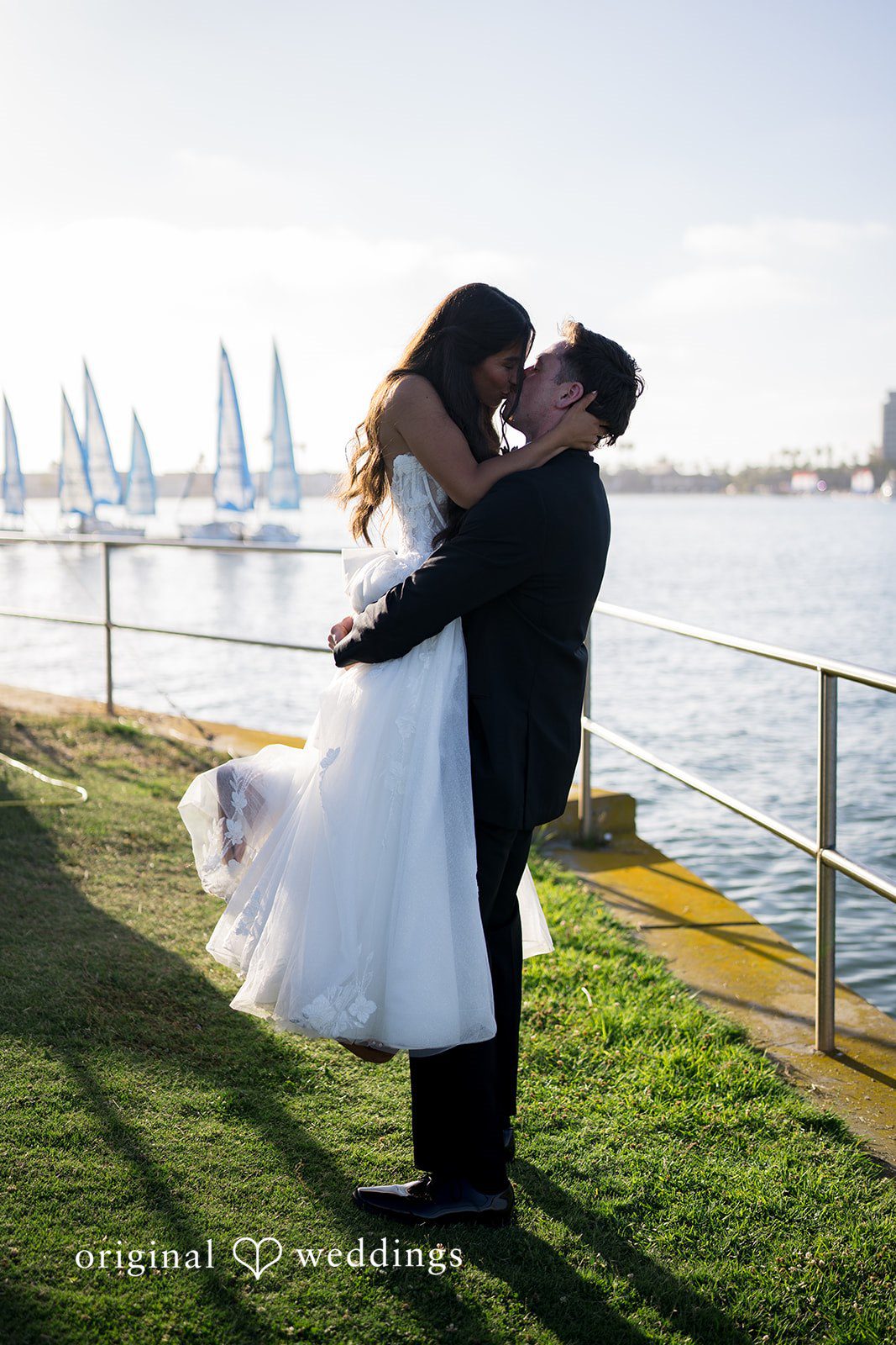 Kalina + Malakai Groom lifting the bride into the air for a kiss next to a waterfront with sailboats.
