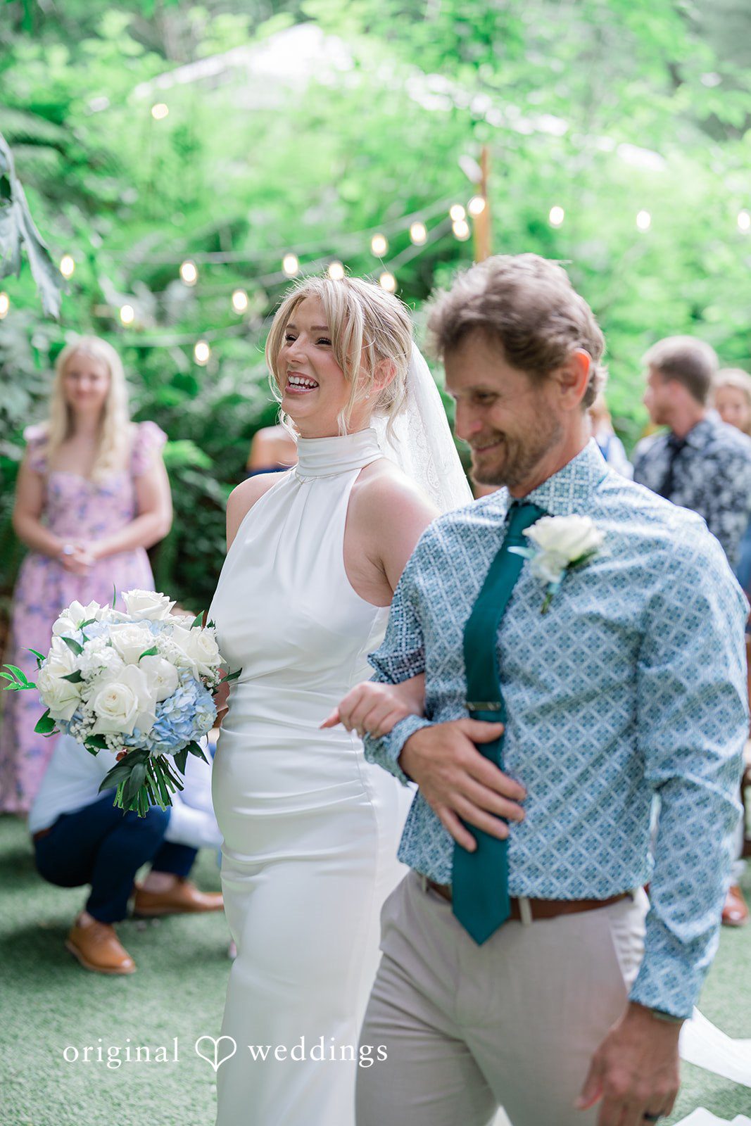 Destiny + Maksim A bride and a man in a striped shirt and blue tie walking together through a garden.