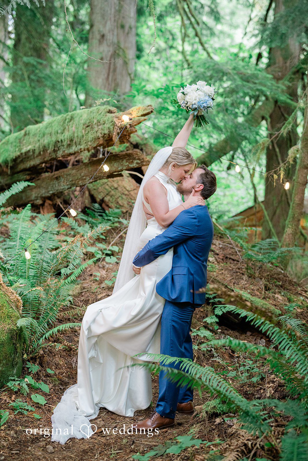 Destiny + Maksim A bride and groom embracing and kissing against a fallen tree trunk in the woods.