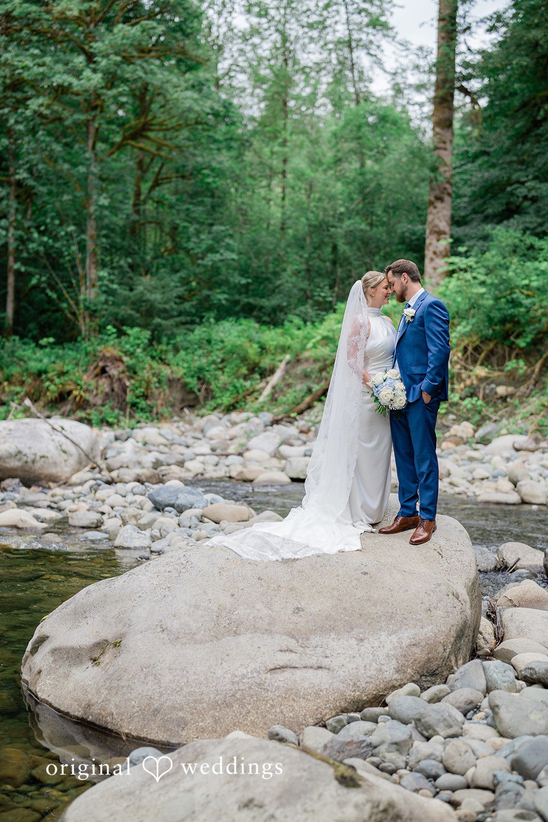Destiny + Maksim A bride and groom posing together on a large boulder by a shallow stream.