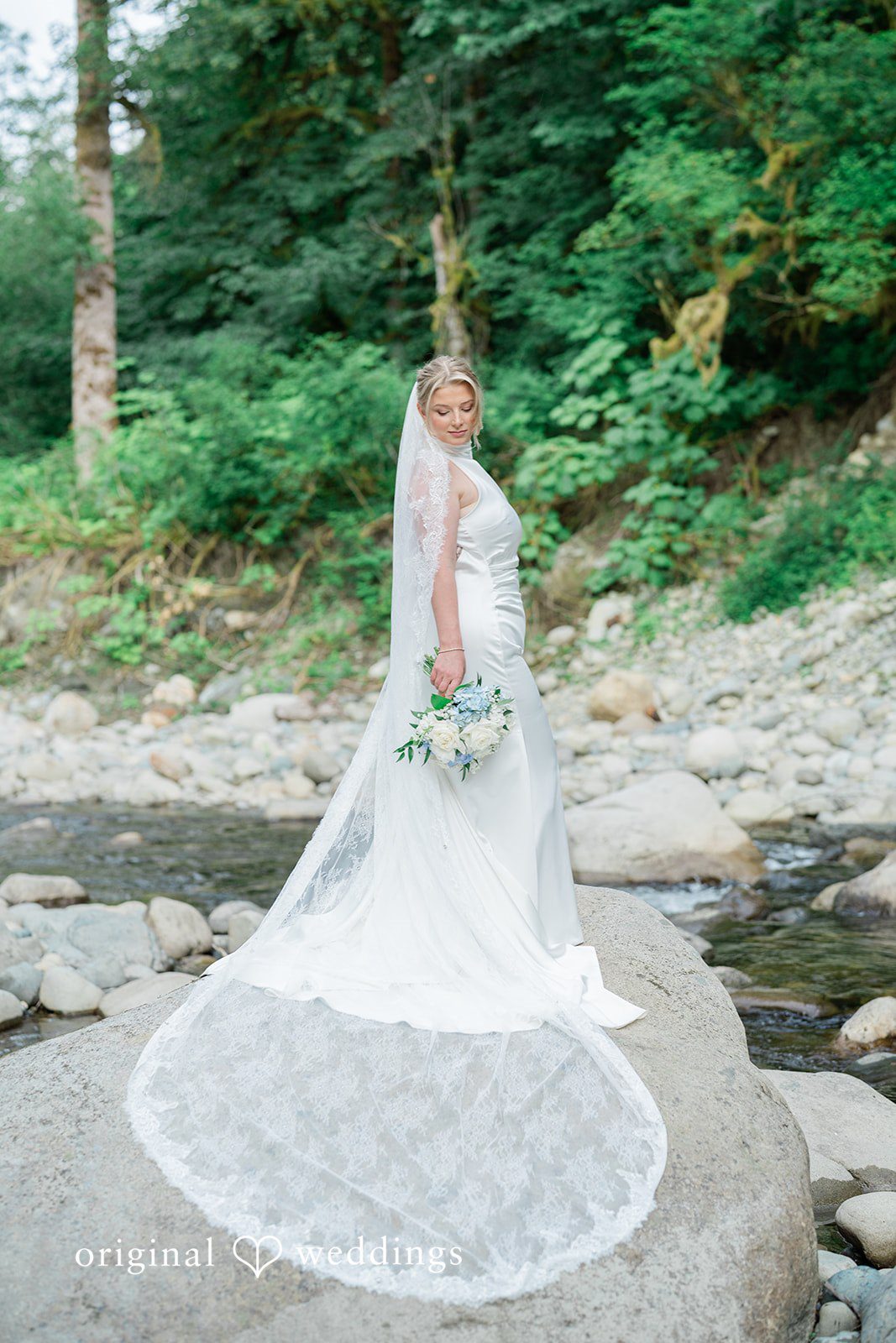 Destiny + Maksim A bride standing on a rock by a stream with her long lace veil trailing behind her.