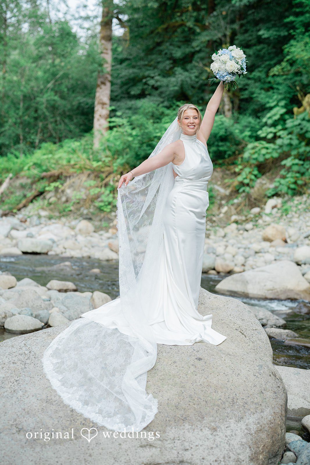 Destiny + Maksim A bride holding her veil up while standing on a large rock by a stream.
