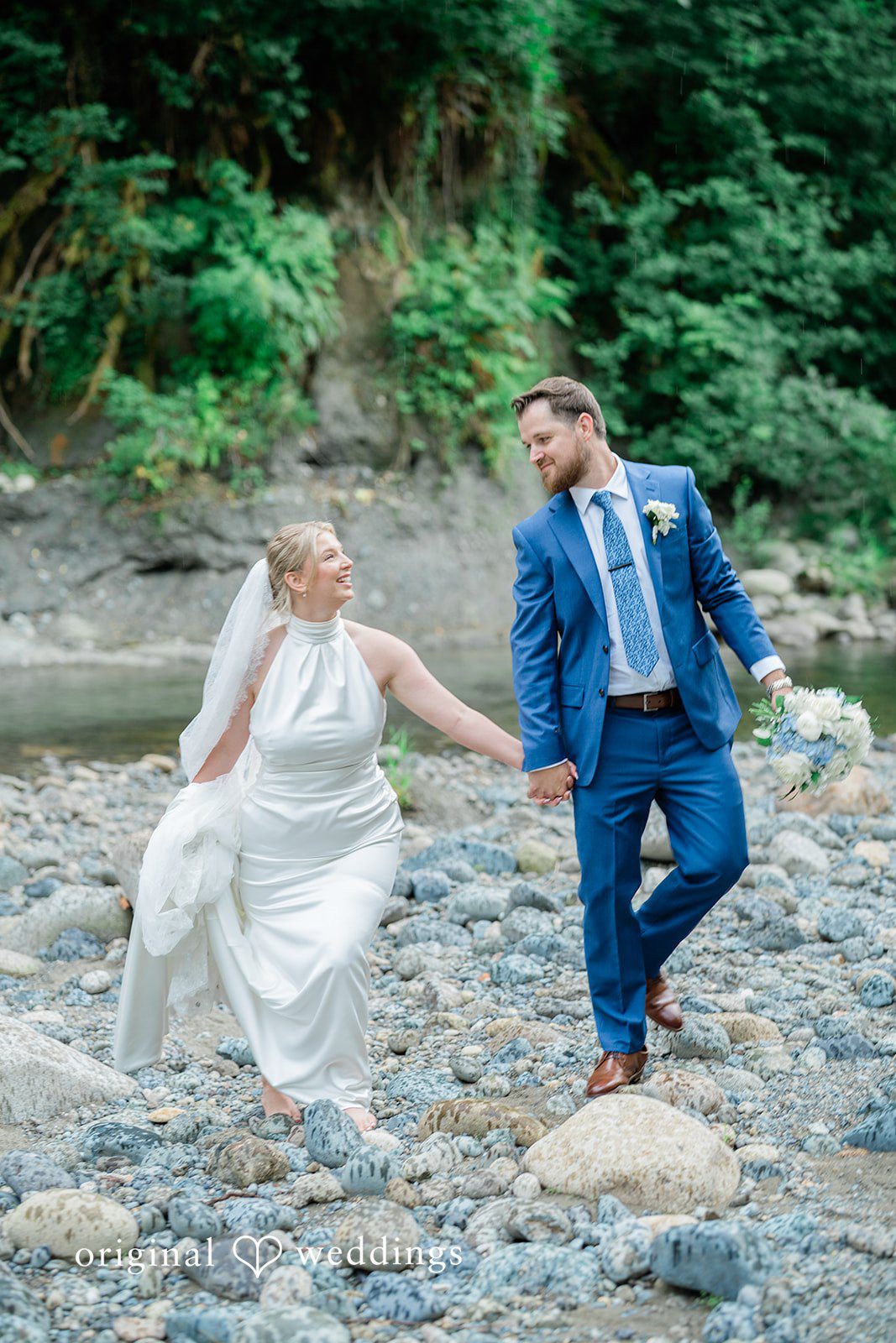 Destiny + Maksim A bride and groom holding hands while stepping across rocks in a shallow river.