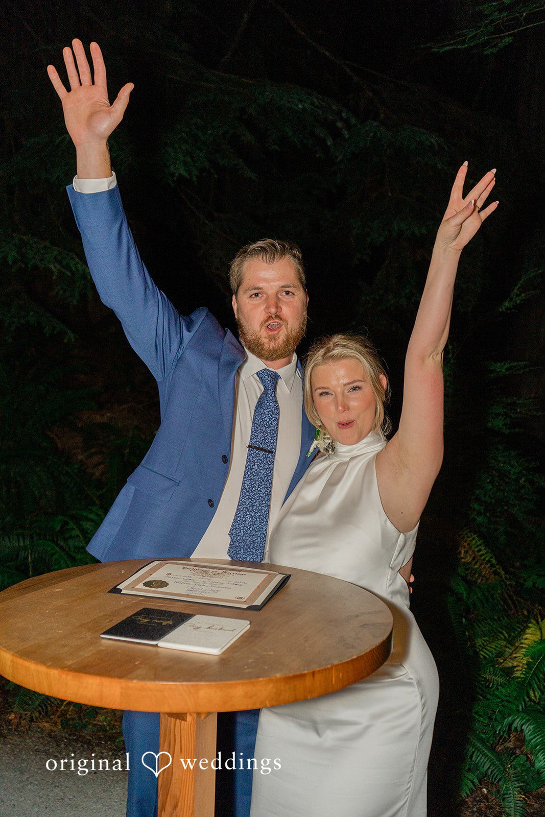 Destiny + Maksim A bride and groom sitting at a table with their arms raised in mid-celebration.