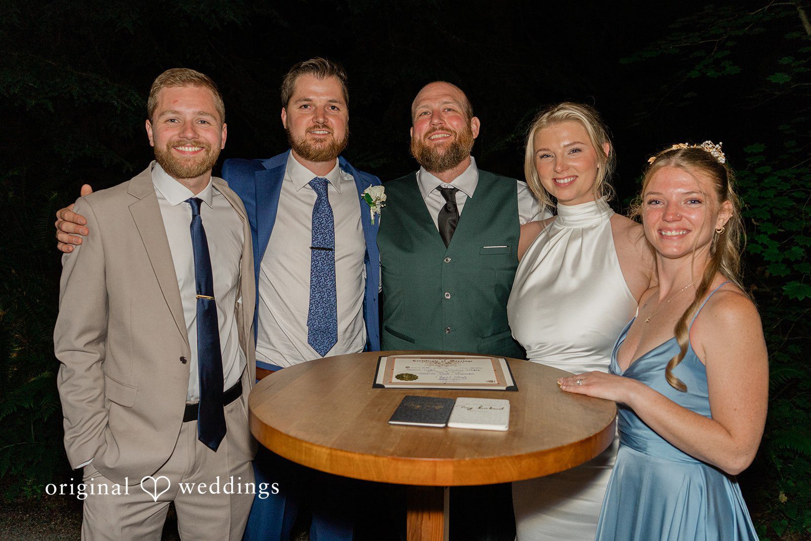 Destiny + Maksim A bride and groom posing for a group photo with wedding guests around a tall table.