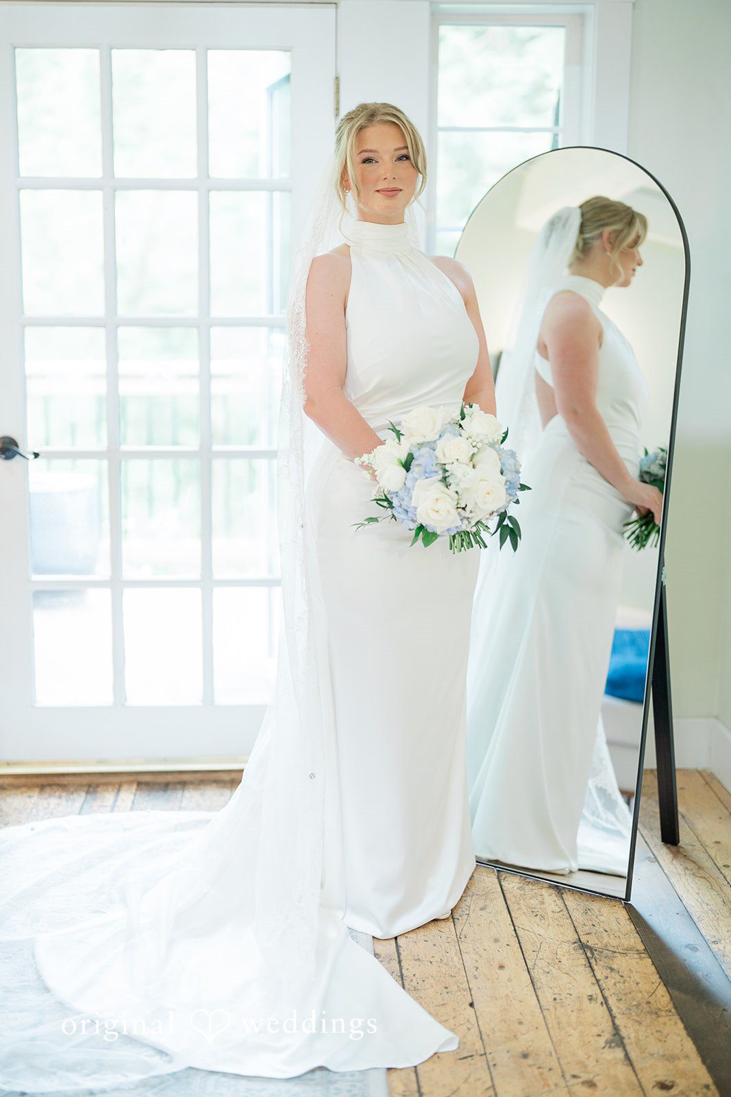 Destiny + Maksim A bride in a sleeveless white gown looking at her reflection in a large mirror.