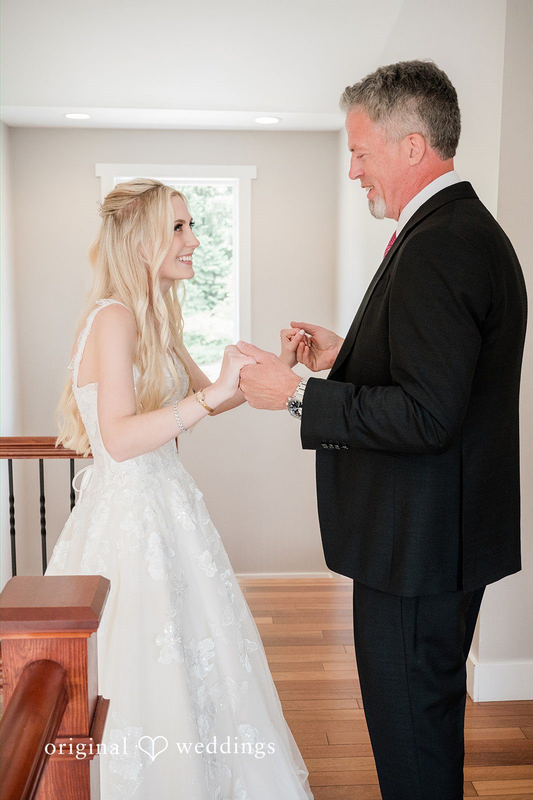 Luke + Kelsey A bride smiles warmly while holding hands with a man wearing a black suit on a wooden floor near a staircase.
