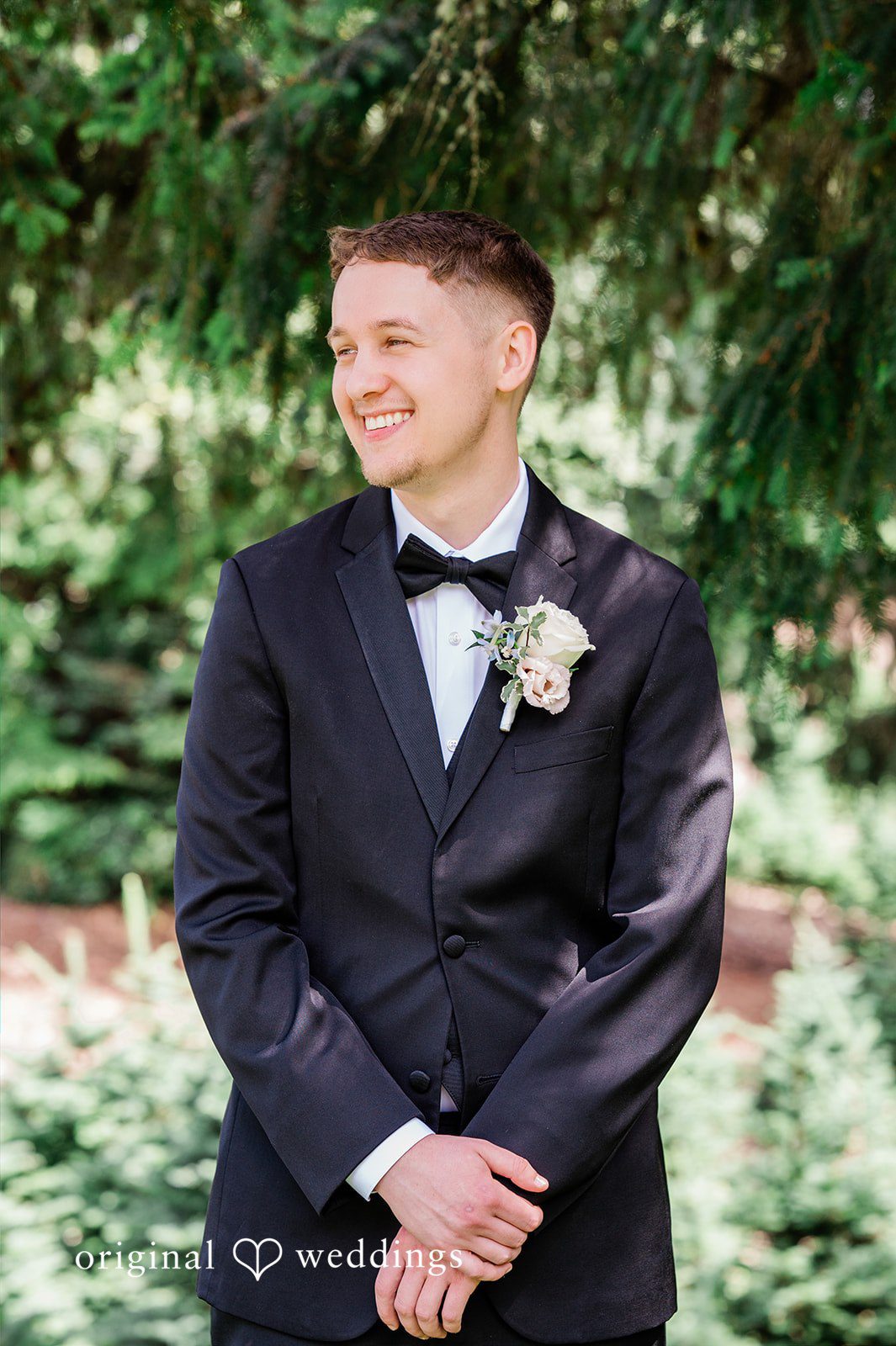 Luke + Kelsey A groom posing solo in a dark tuxedo with a bowtie against a backdrop of trees.