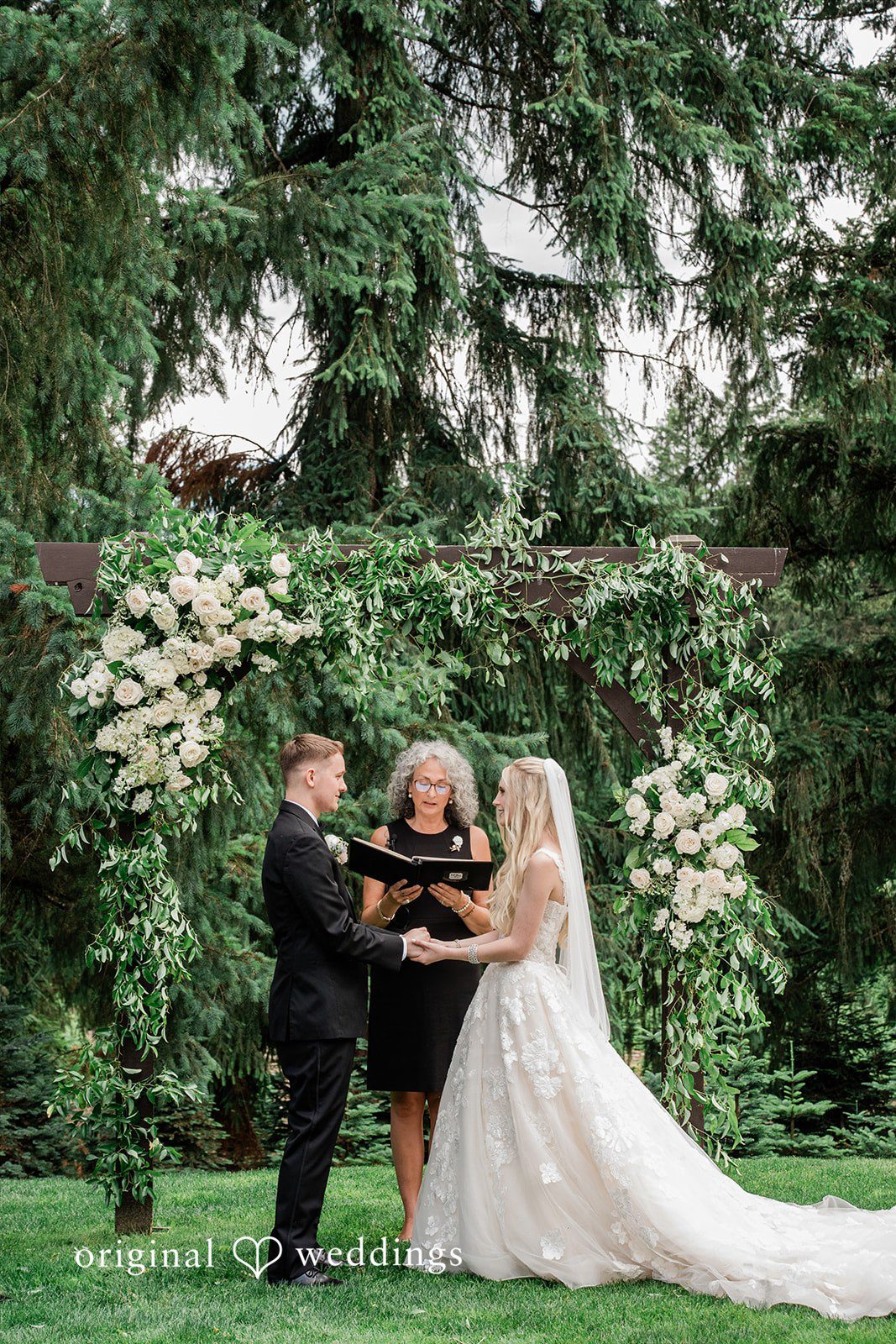 Luke + Kelsey A wedding ceremony taking place in front of an officiant and a greenery arch.