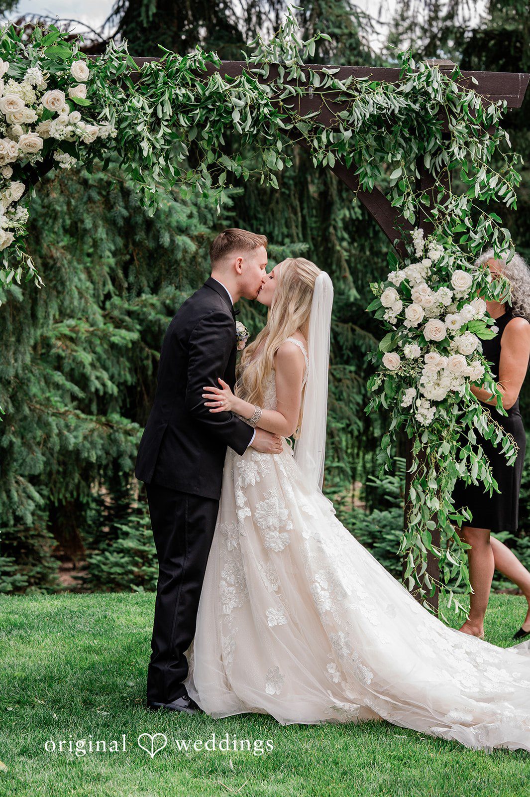 Luke + Kelsey A bride and groom sharing a kiss against a backdrop of dark green trees.