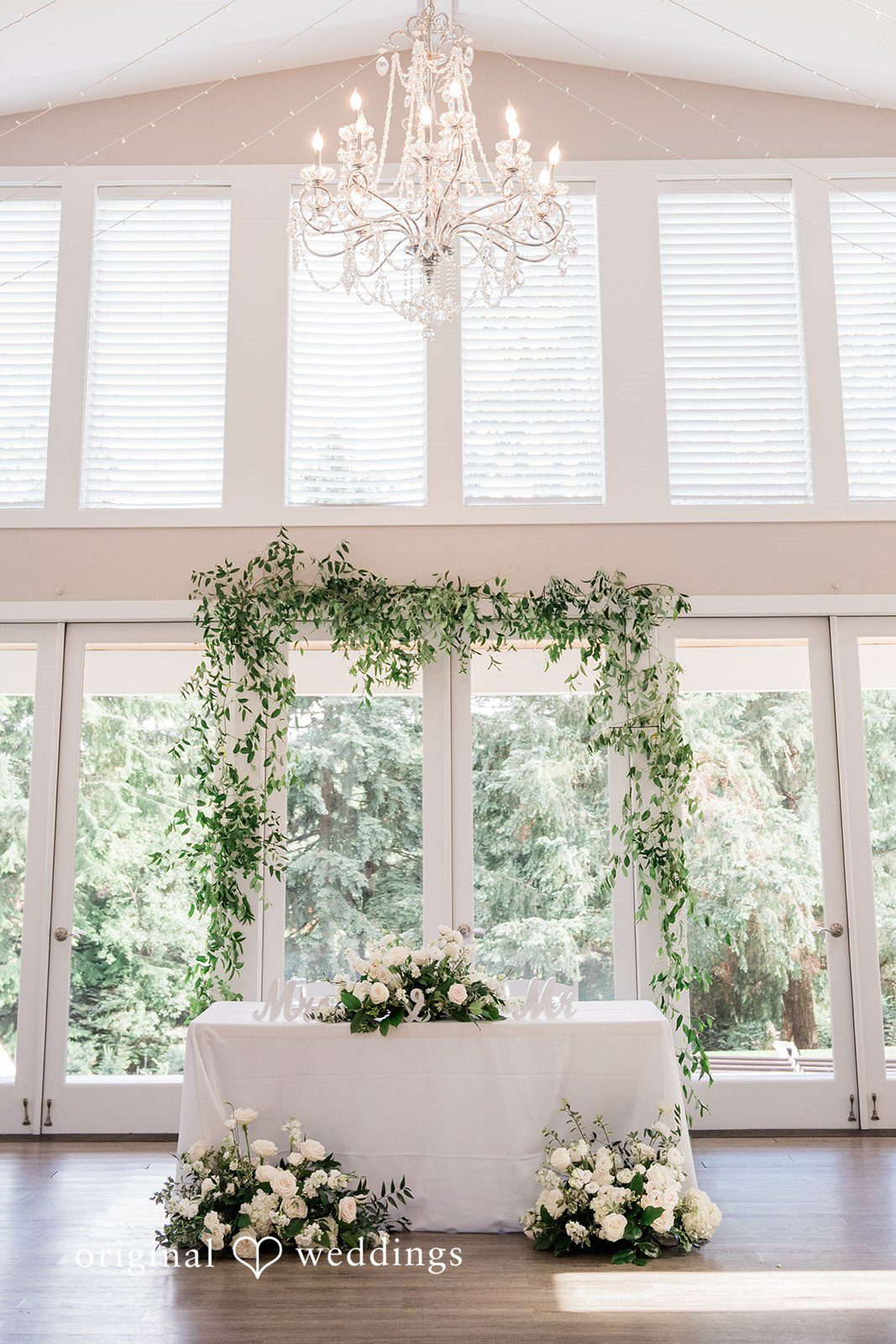 Luke + Kelsey A wedding reception head table decorated with a large floral and greenery arch.