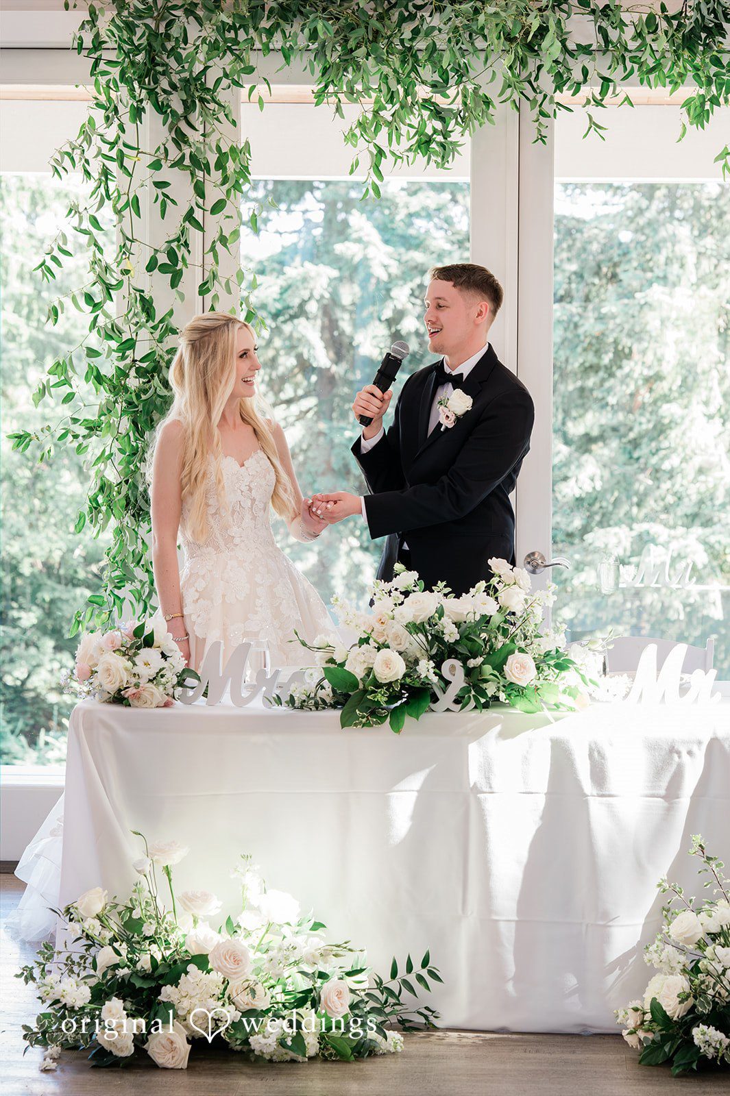 Luke + Kelsey A groom speaks into a microphone while holding his bride's hand at a flower-decorated wedding reception table.