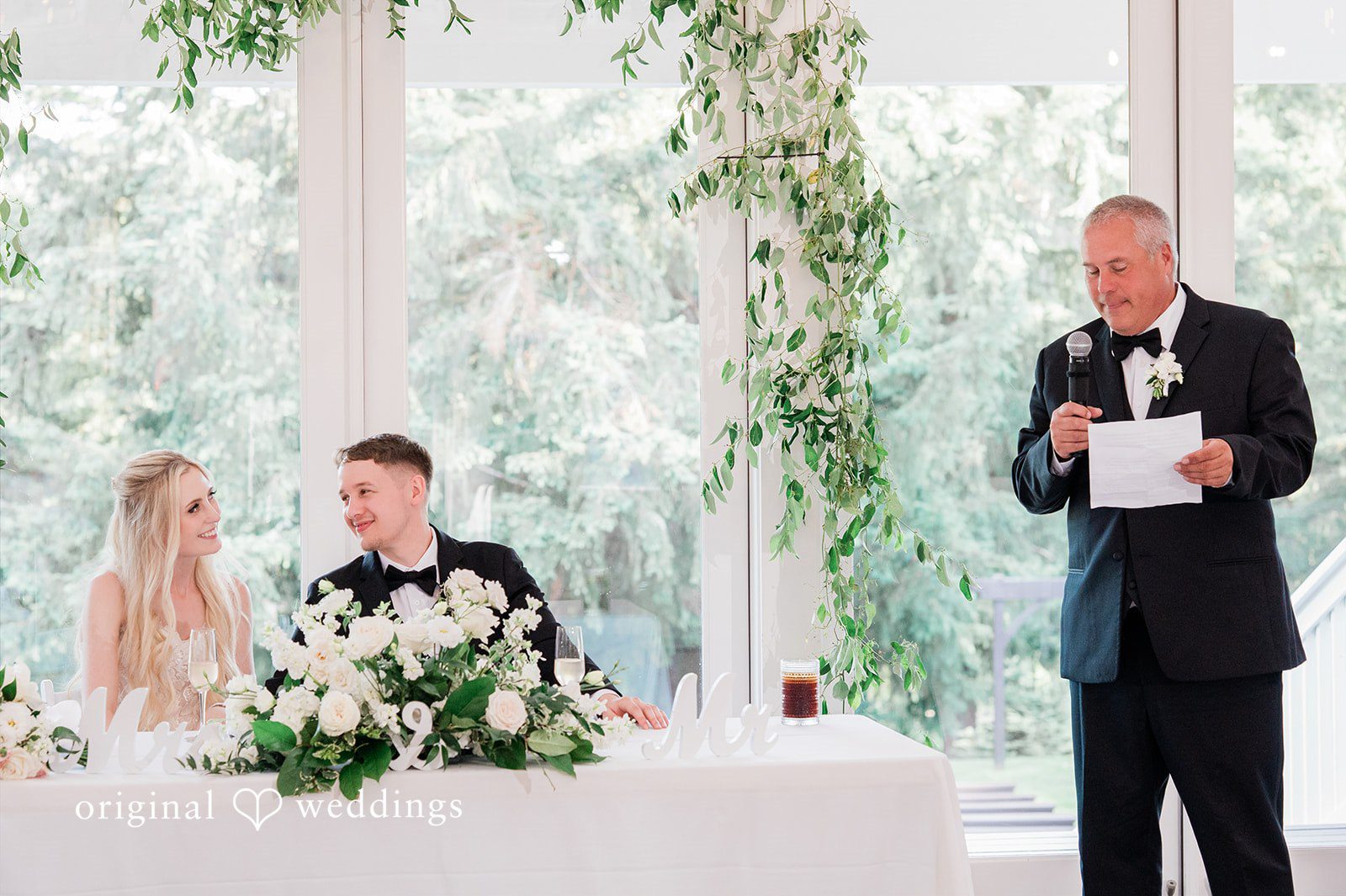 Luke + Kelsey A man speaking into a microphone while a bride and groom listen at a table.