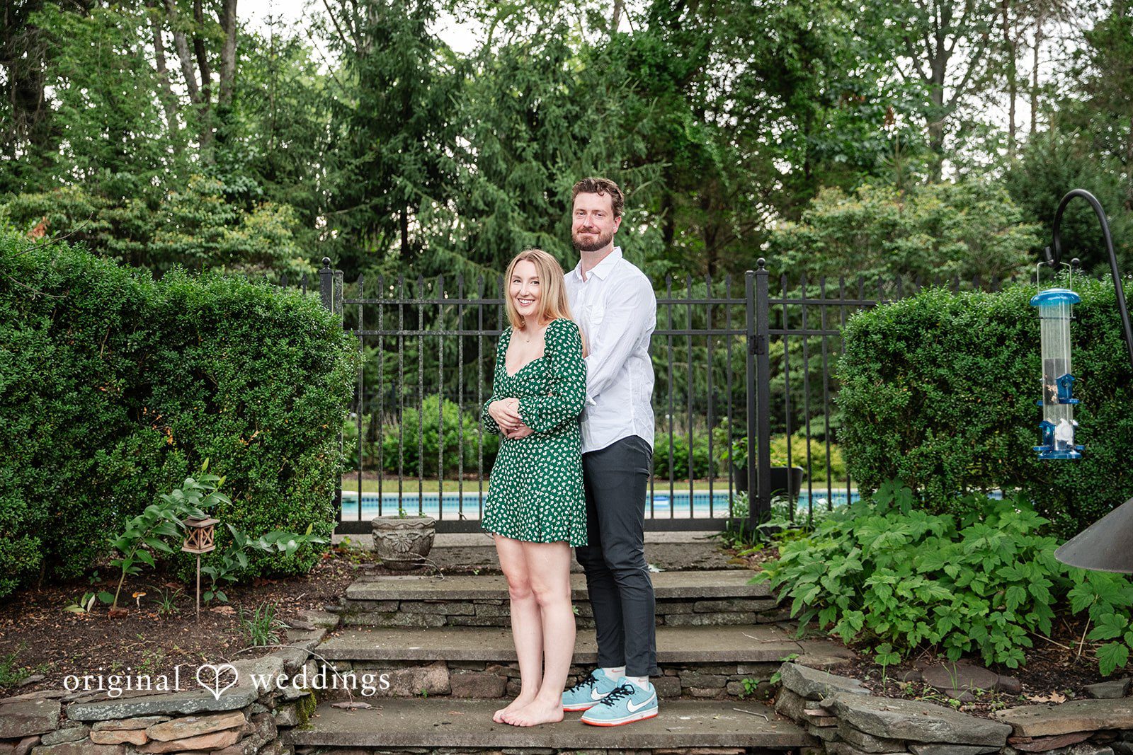 Sarah + Tyler A couple stands together smiling on a stone pathway in a park-like setting.