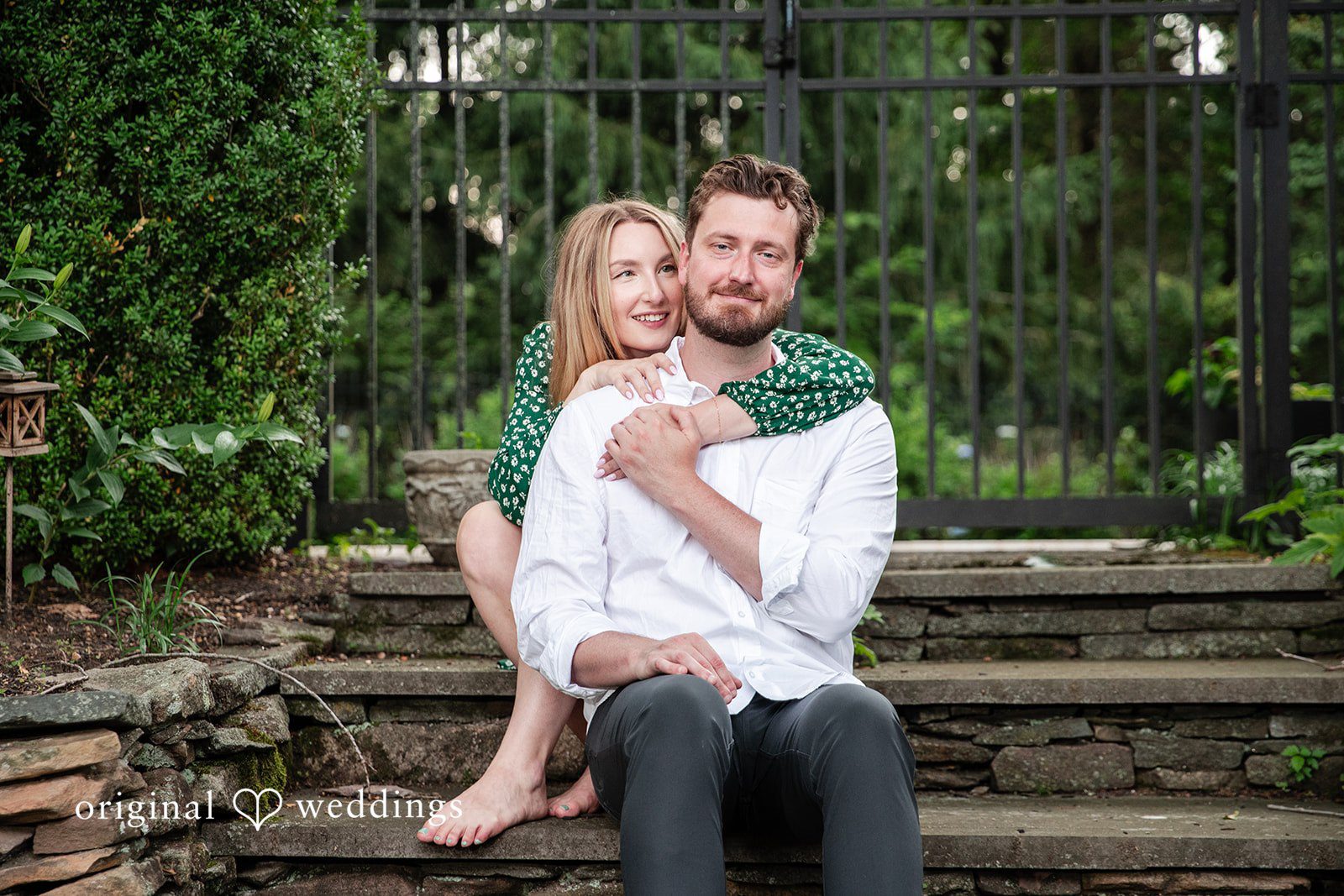 Sarah + Tyler A man holds a woman in his lap while sitting on stone steps surrounded by greenery.