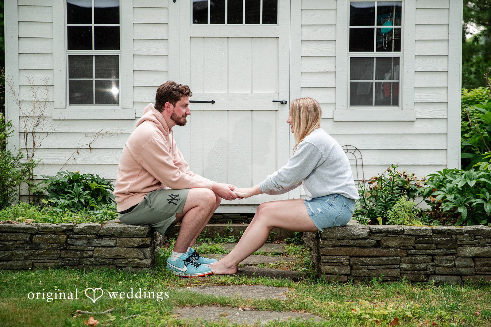 Sarah + Tyler A couple sits on a stone wall holding hands in front of a white building.
