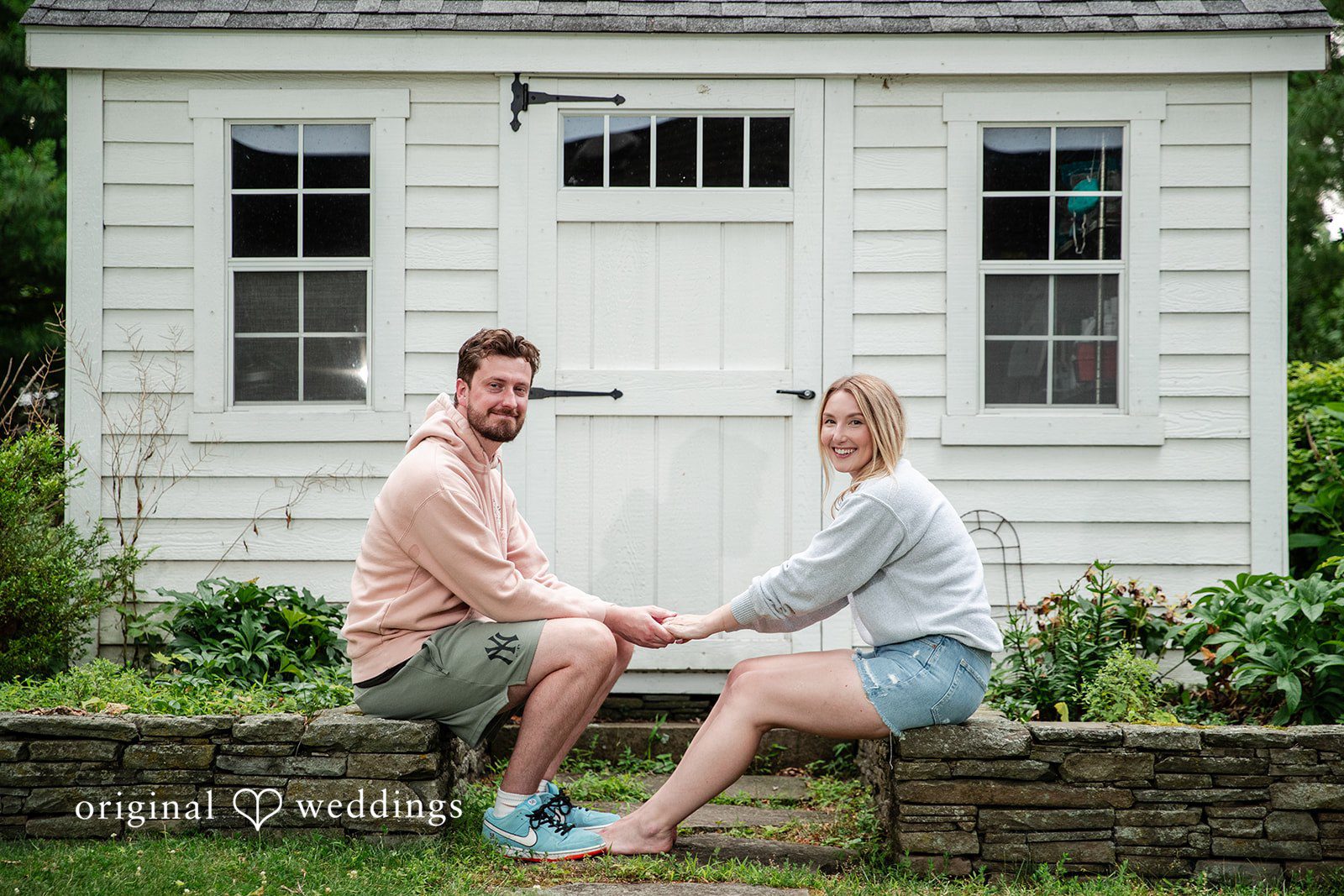 Sarah + Tyler A couple sits on a low stone wall chatting in front of a white house.