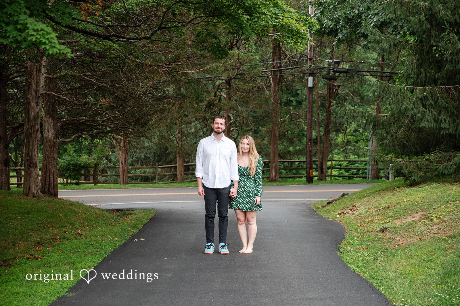 Sarah + Tyler A couple walks hand-in-hand down a paved path surrounded by lush green trees.