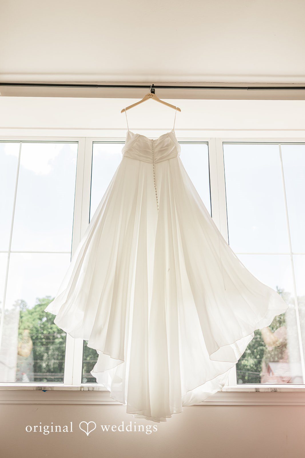 Emily + Joshua Elegant wedding dress hanging in front of a sunlit window before the ceremony.