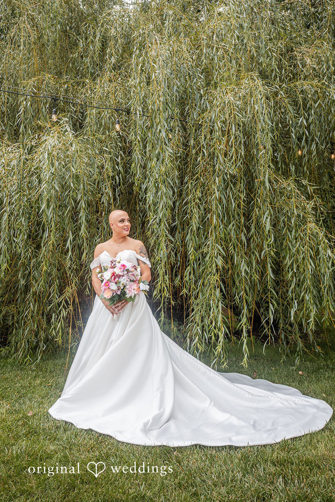 Savannah + Phillip A bride in a white wedding gown with a long train posing on a lawn near a willow tree.