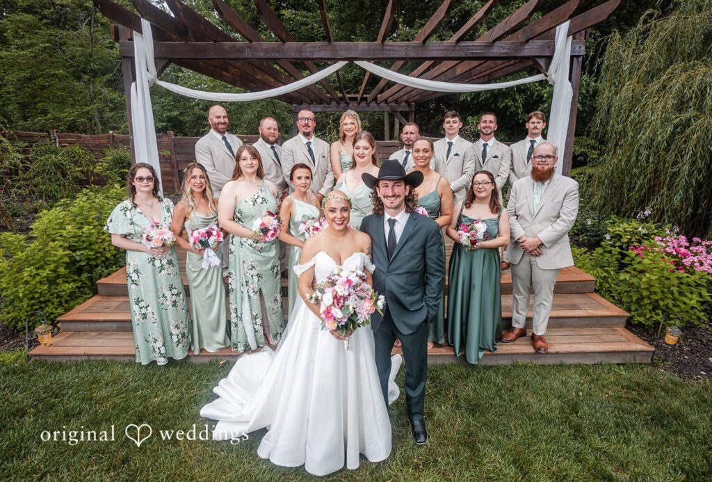 Savannah + Phillip A full wedding party posing together on a grassy lawn in front of a rustic altar structure at Willow Acres captured by Indianapolis wedding photographers from Original Weddings