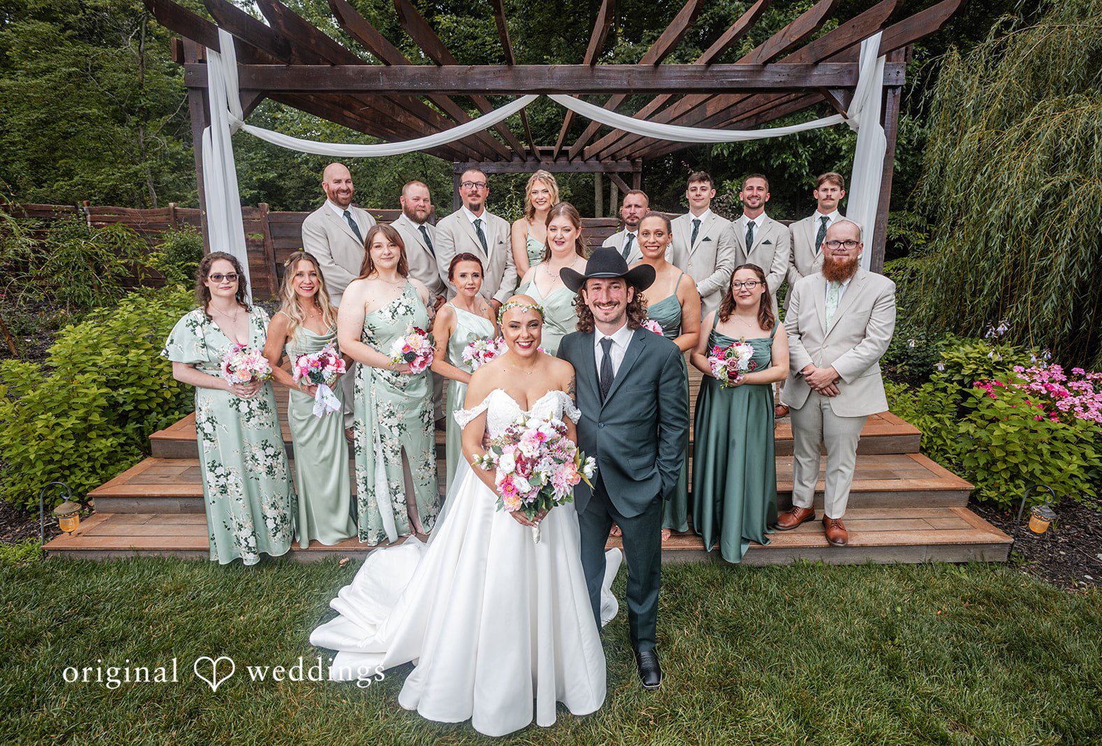A full wedding party posing together on a grassy lawn in front of a rustic altar structure at Willow Acres captured by Indianapolis wedding photographers from Original Weddings