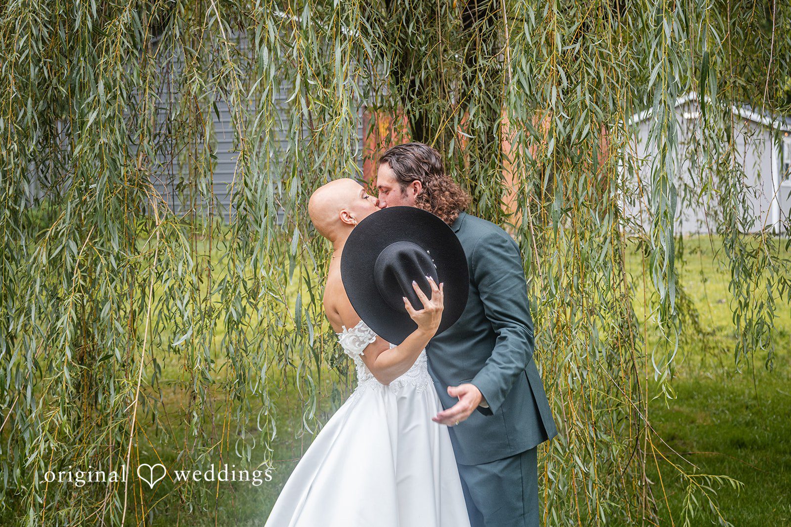 Savannah + Phillip A bride and a groom in a black cowboy hat embracing in front of a green willow tree backdrop.
