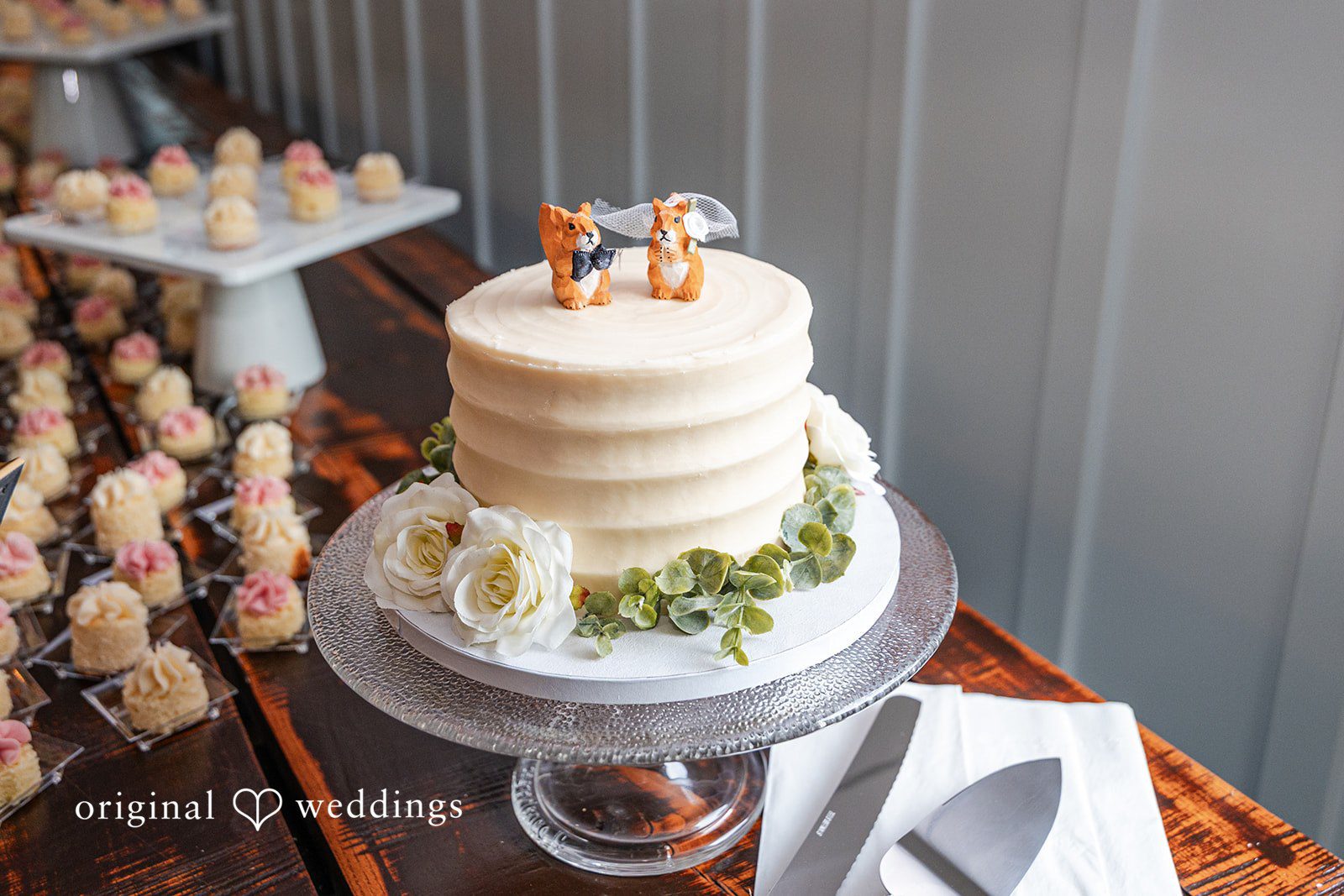Savannah + Phillip A two-tiered white wedding cake decorated with green foliage on a silver stand.