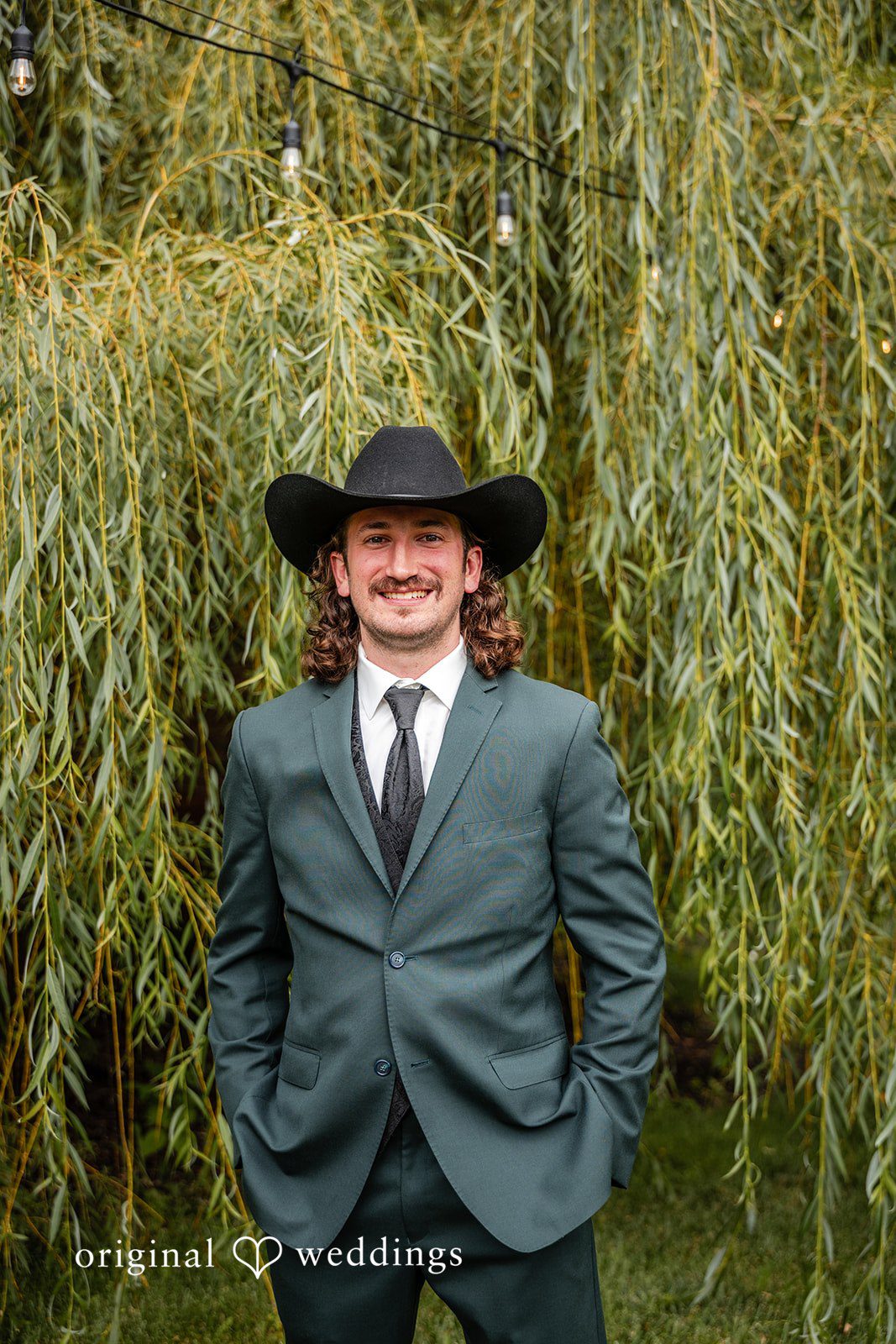 Savannah + Phillip A groom wearing a dark suit and a black cowboy hat smiling in front of a willow tree.