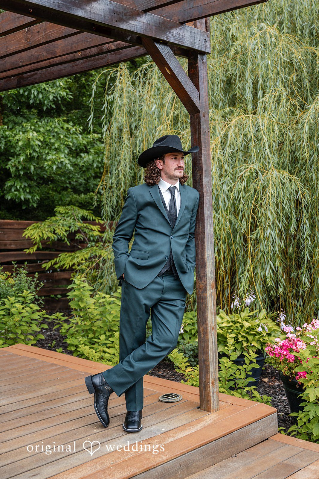 Savannah + Phillip A groom in a dark suit and black cowboy hat leaning casually against a wooden railing.