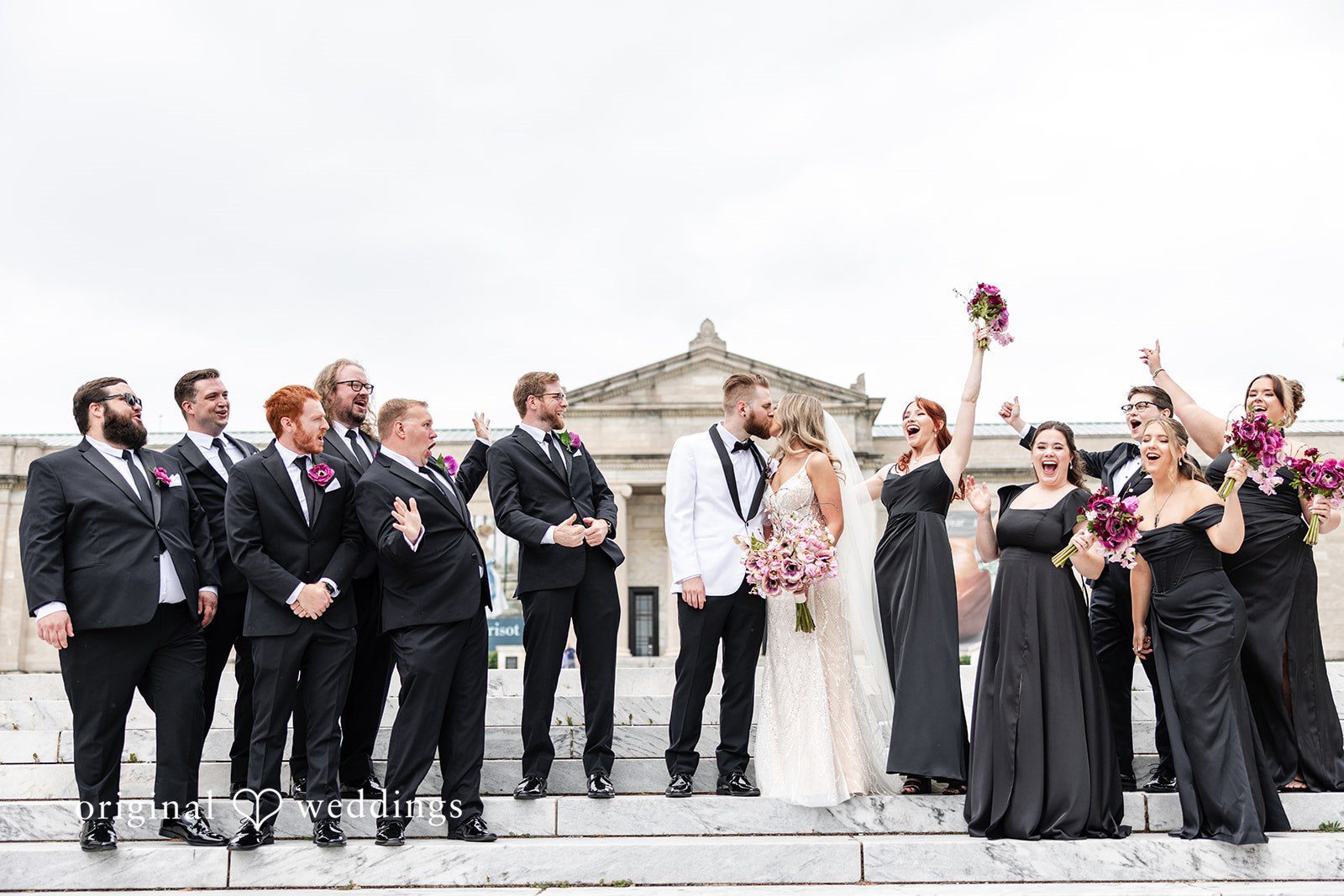Dan + Alex A wedding party cheering and raising their hands together on wide stone stairs.