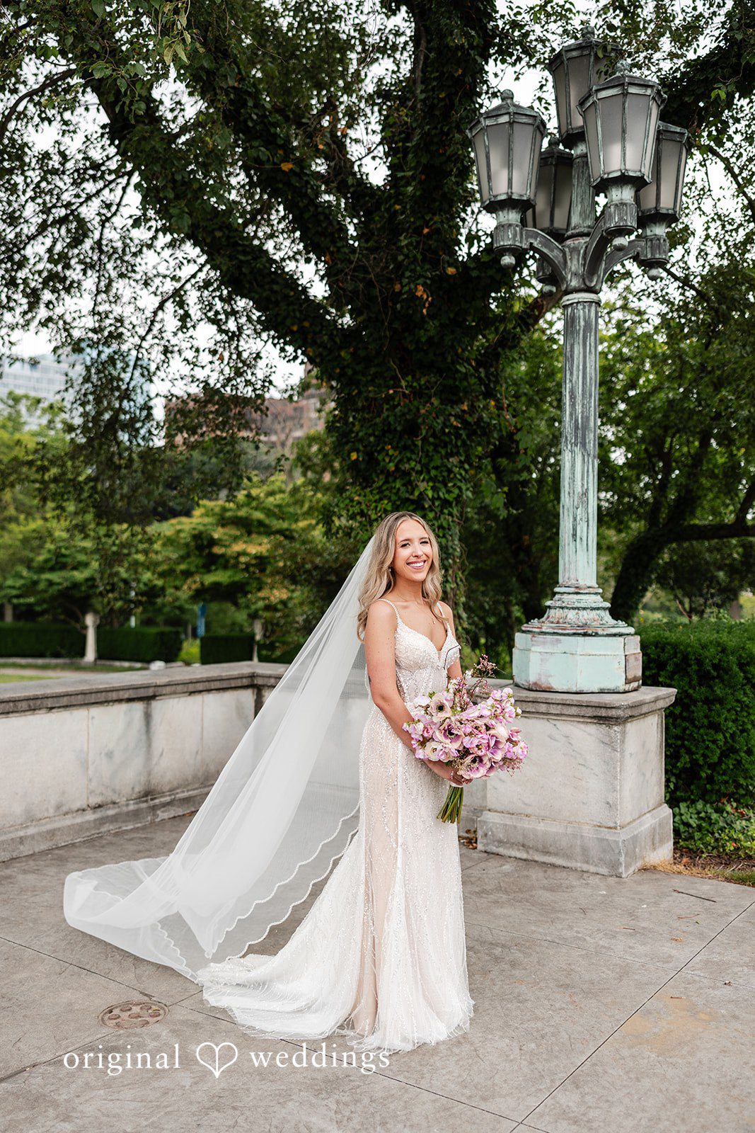 Dan + Alex A bride in a lace gown and long veil posing on a pathway with lush green trees.