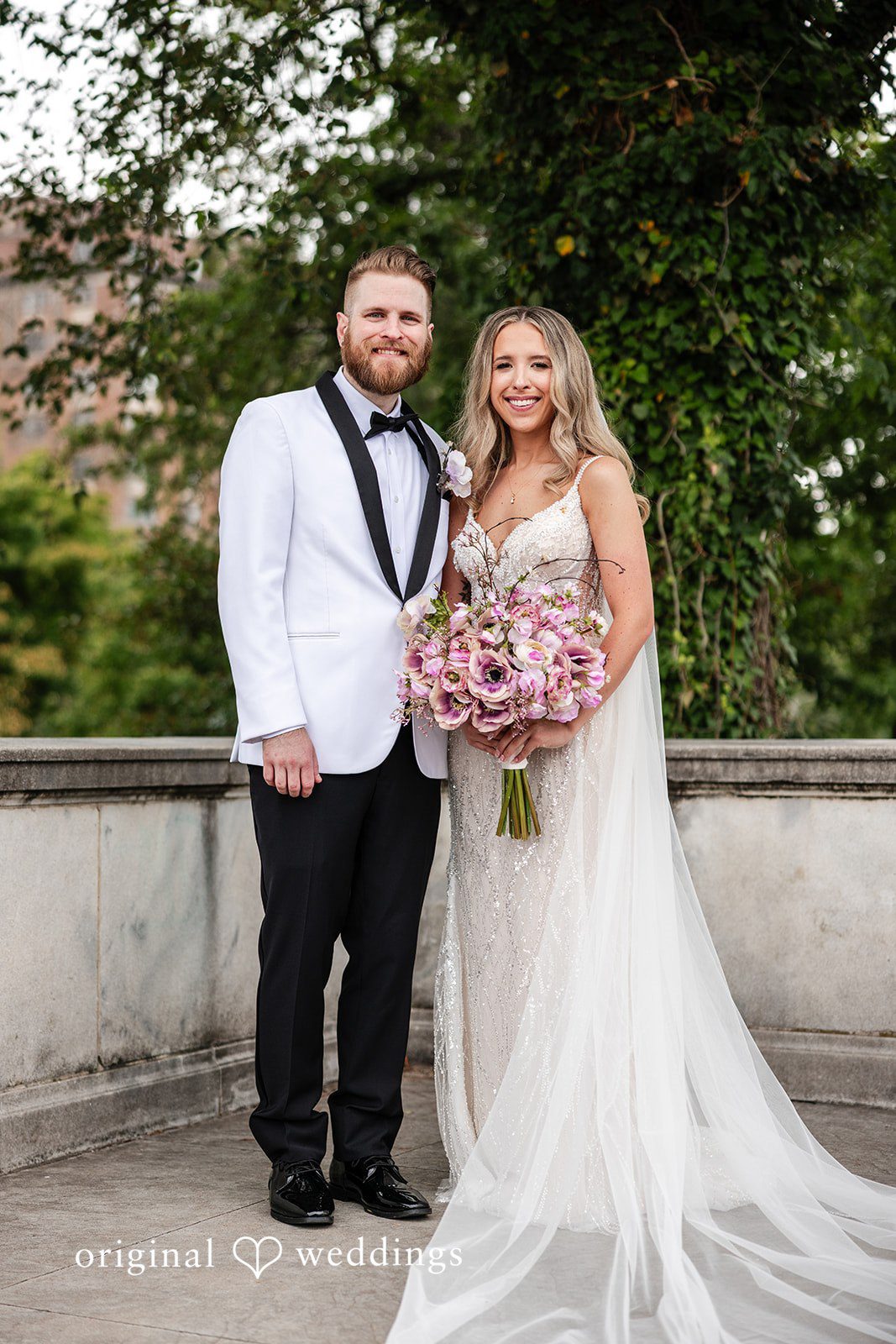 Dan + Alex A bride holding a bouquet and a groom smiling together on a stone balcony with trees behind them.