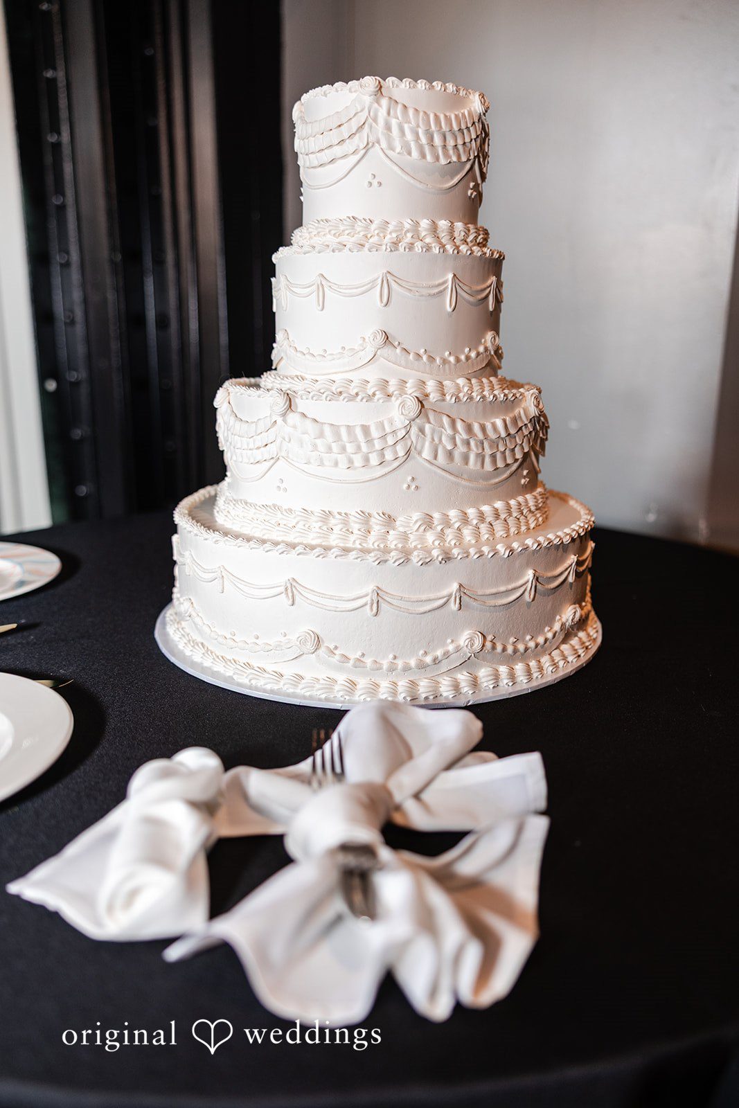 Dan + Alex A multi-tiered white wedding cake decorated with textured frosting on a dark table.