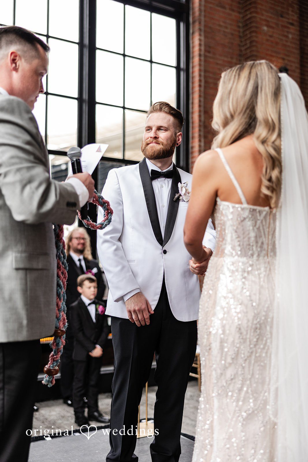 Dan + Alex A bride in an embellished gown and a groom in a white jacket standing together at the altar.