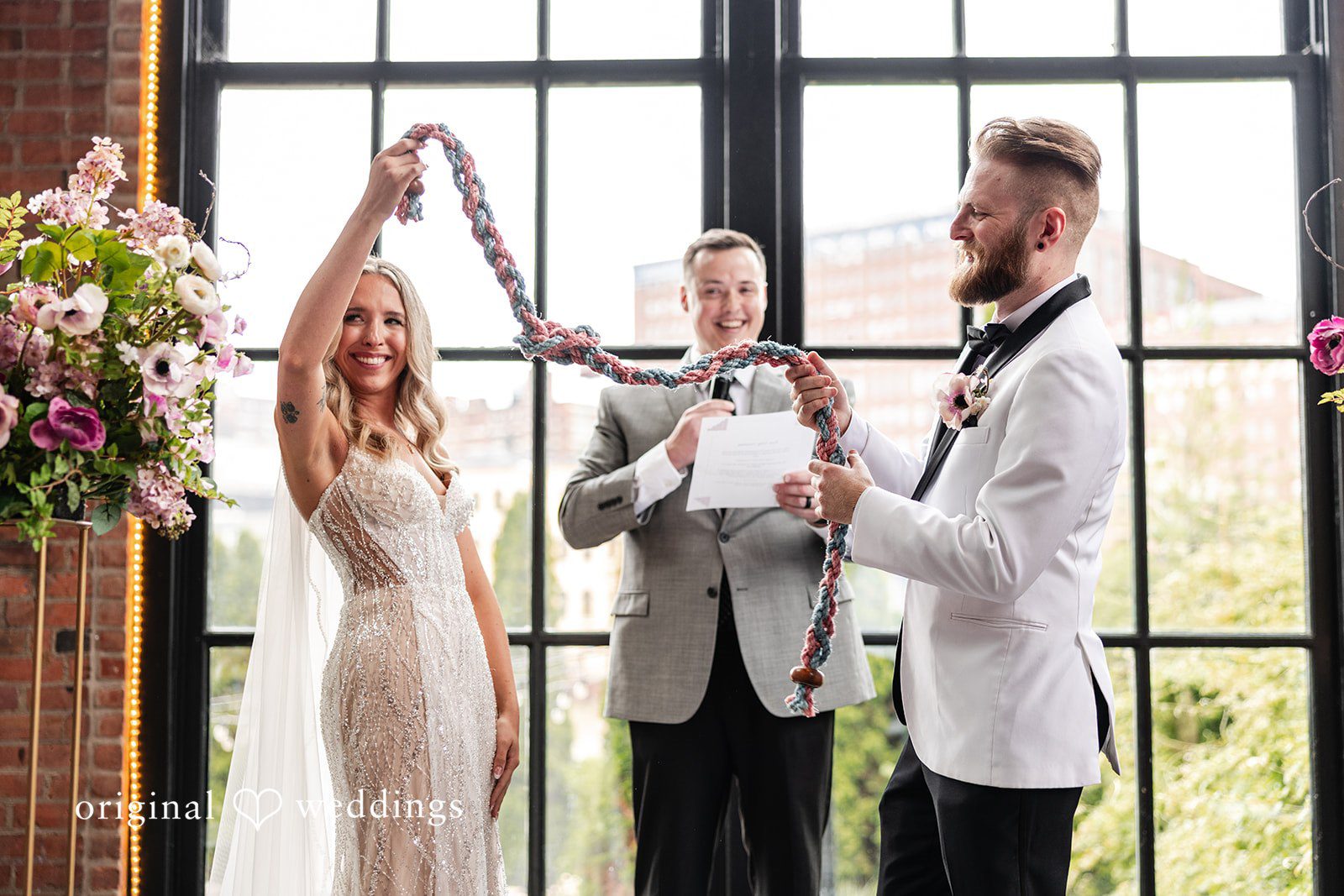 Dan + Alex A wedding officiant speaking to a bride and groom during a ceremony in front of windows.