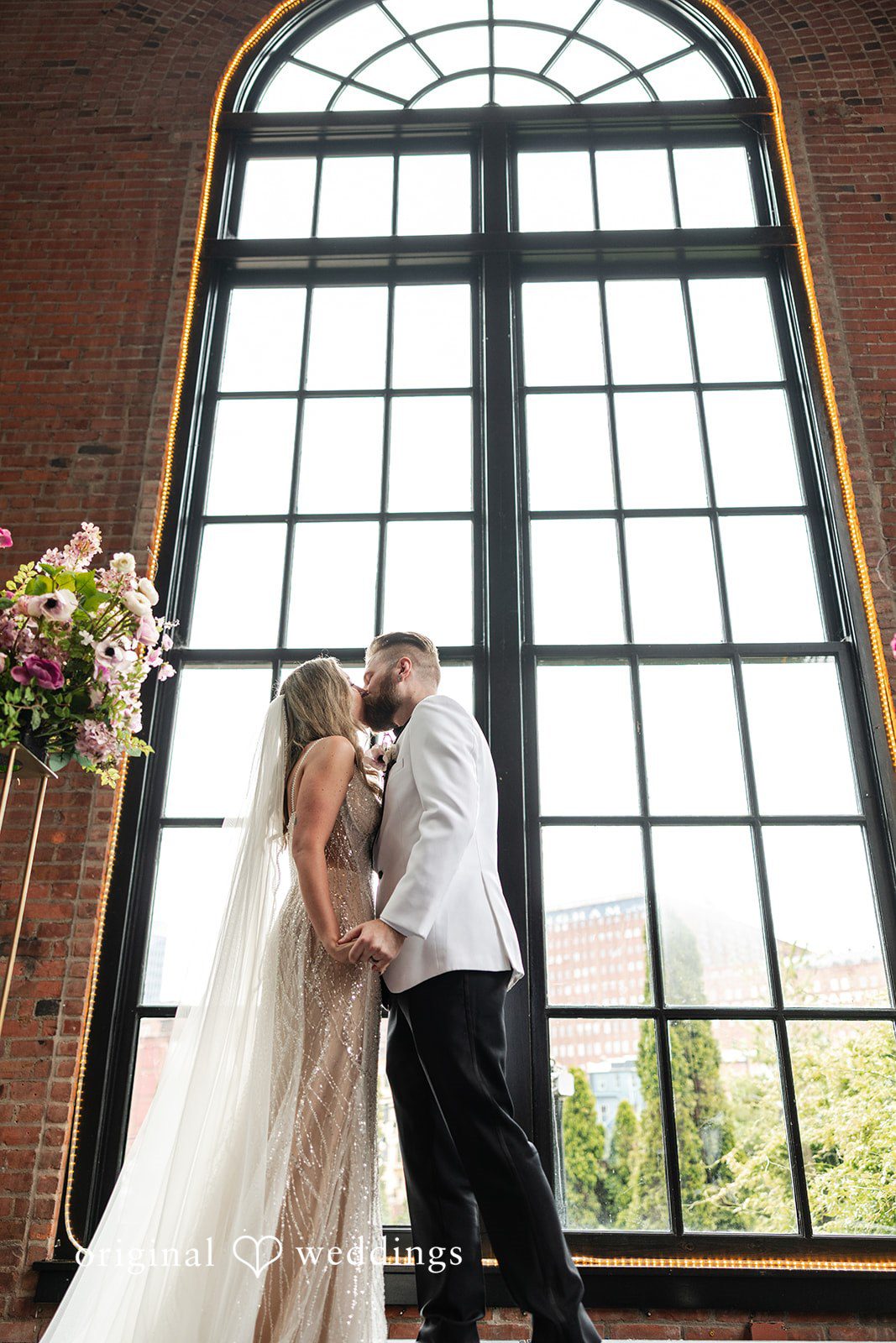 Dan + Alex A bride and groom embracing and kissing in front of a towering arched glass window