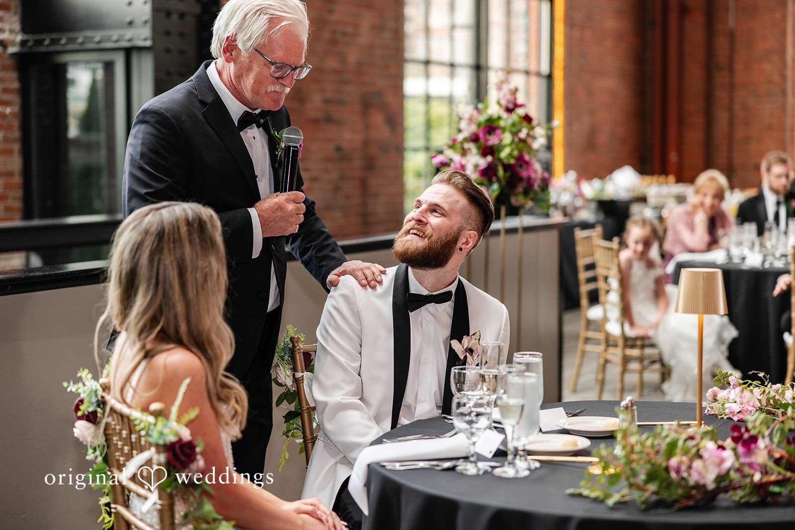 Dan + Alex A groom sitting at a reception table looking up and chatting with wedding guests.